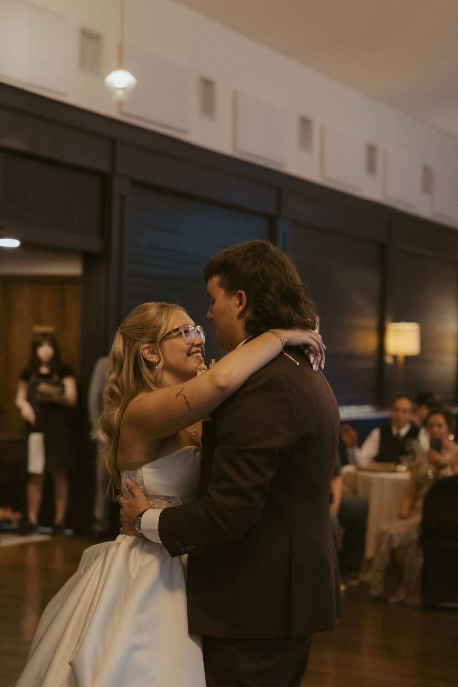 Bride and groom share first dance, smiling at each other in warmly lit wedding reception venue.