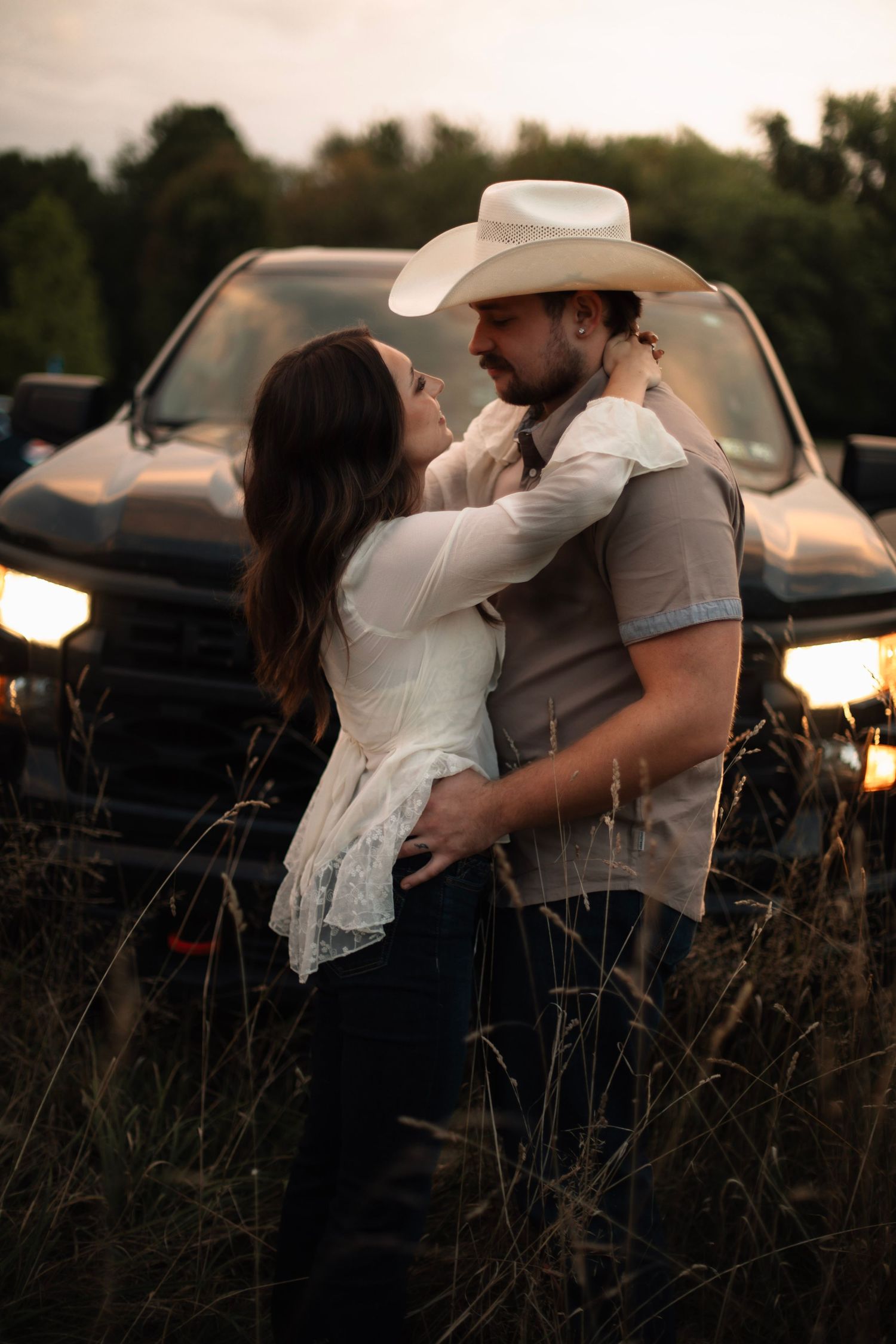 Couple embracing romantically in a field at sunset, man wearing a cowboy hat, with a truck's headlights glowing behind them.