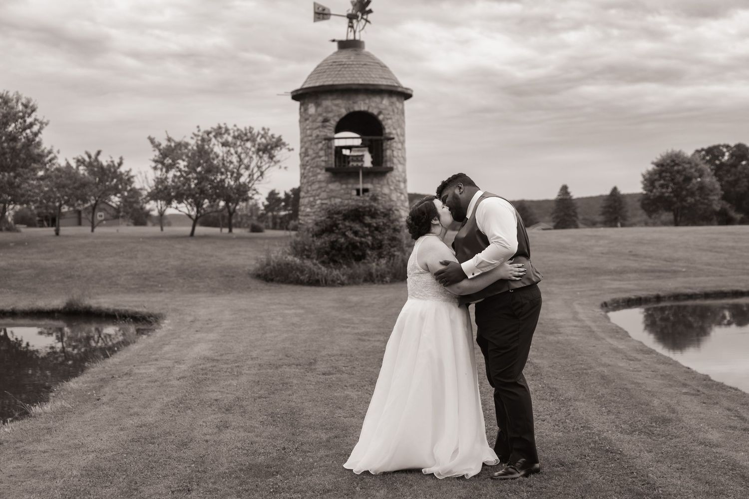 Black and white photo of a bride and groom sharing a romantic moment near a stone windmill and pond on their wedding day.