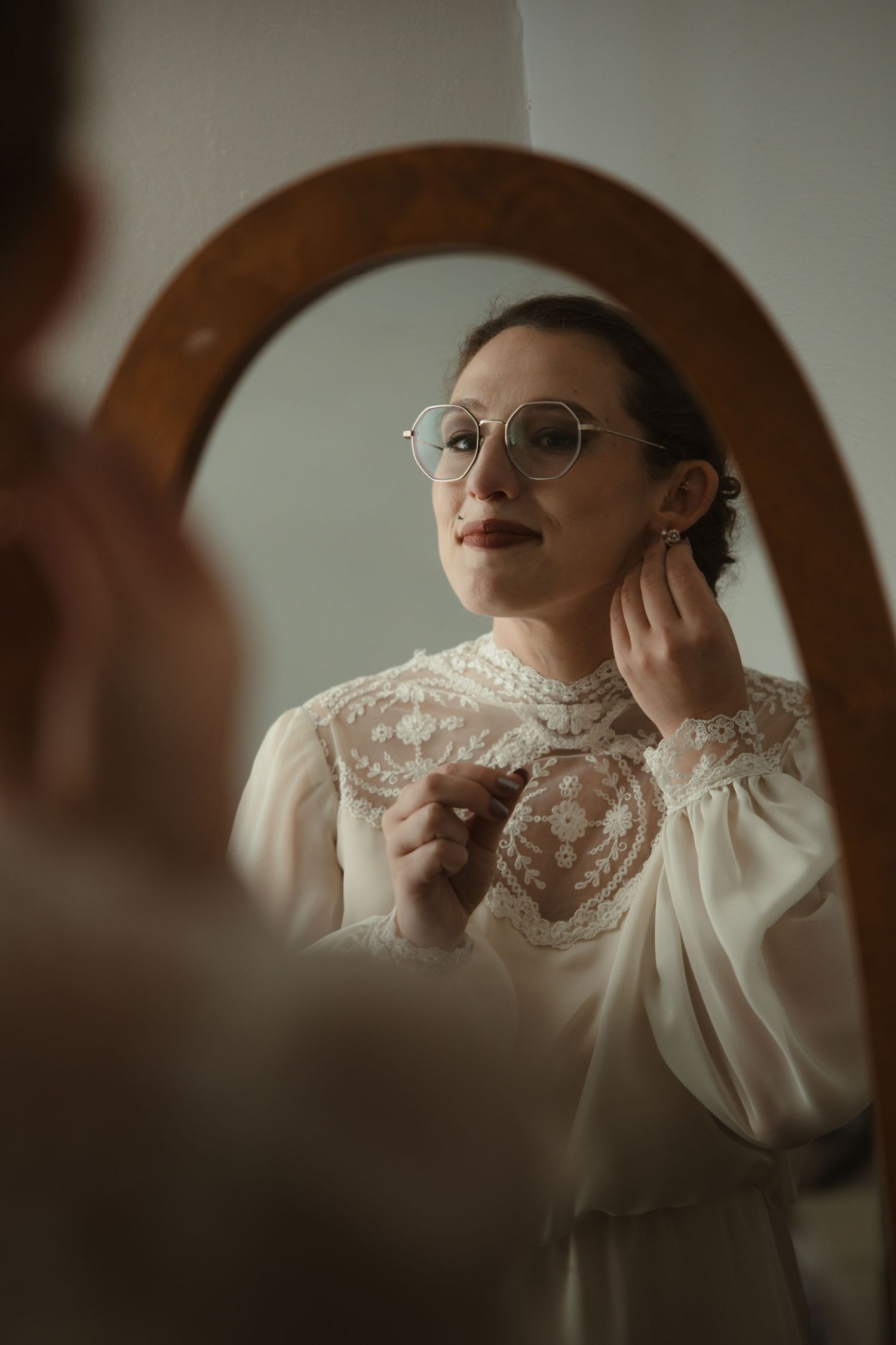 Woman in white lace blouse and round glasses adjusting earring while looking in a wooden framed mirror.