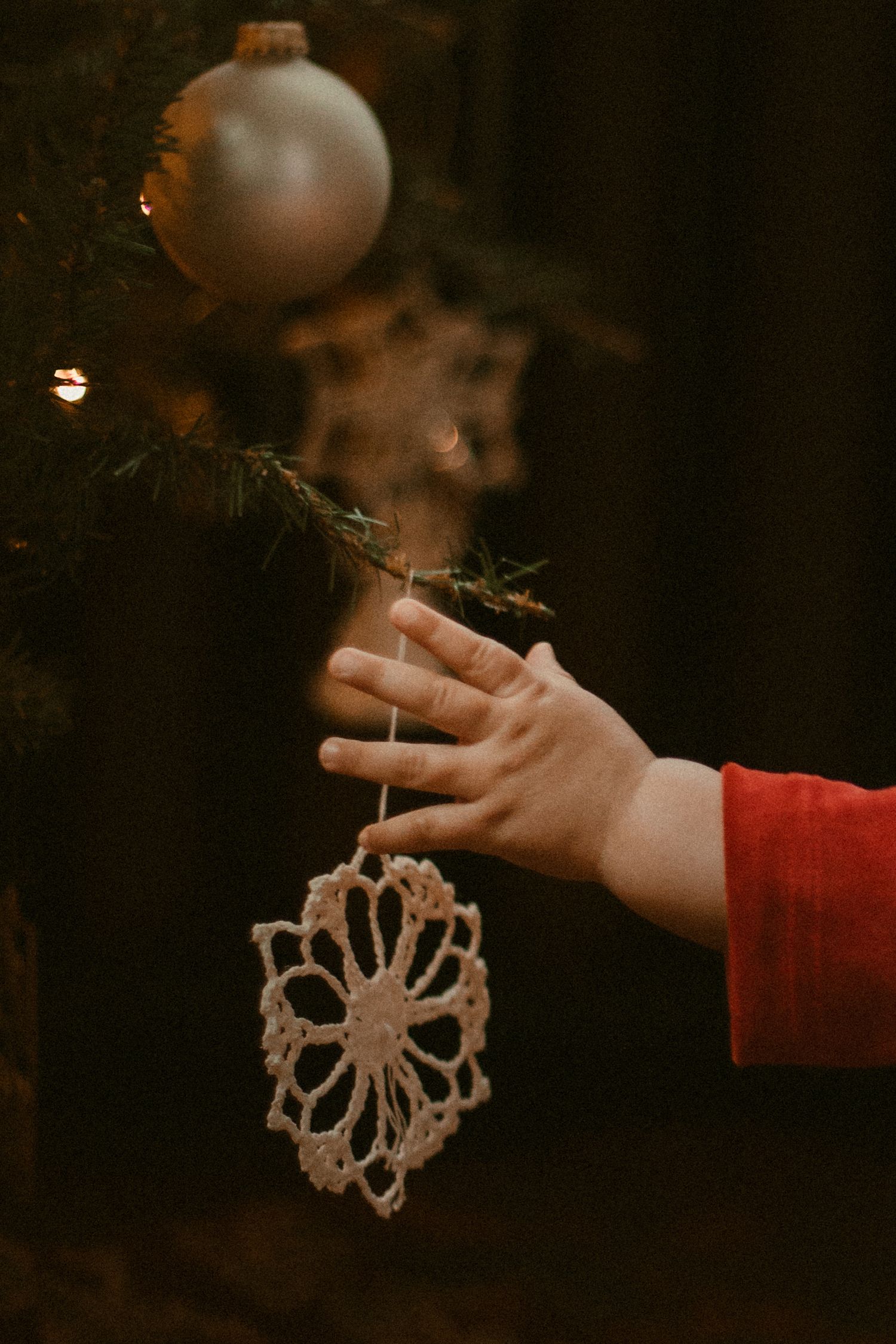 Child's hand hanging a crocheted snowflake ornament on a Christmas tree with a silver ball above.