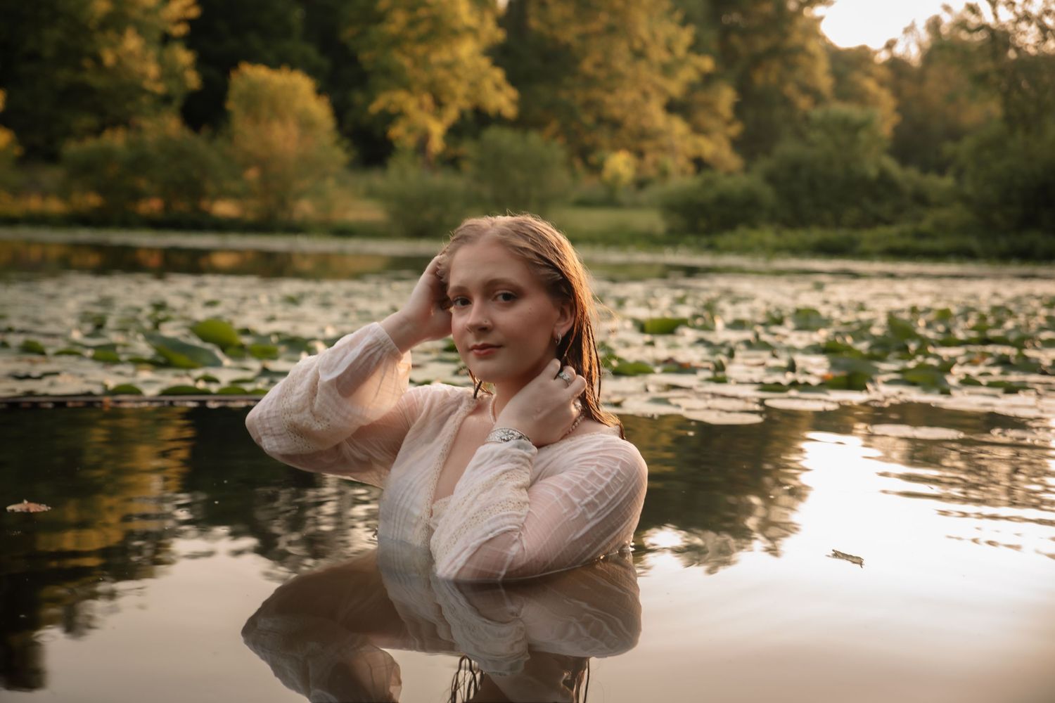 A young woman in a white lace top stands waist-deep in a lily pad-covered pond surrounded by autumn trees.