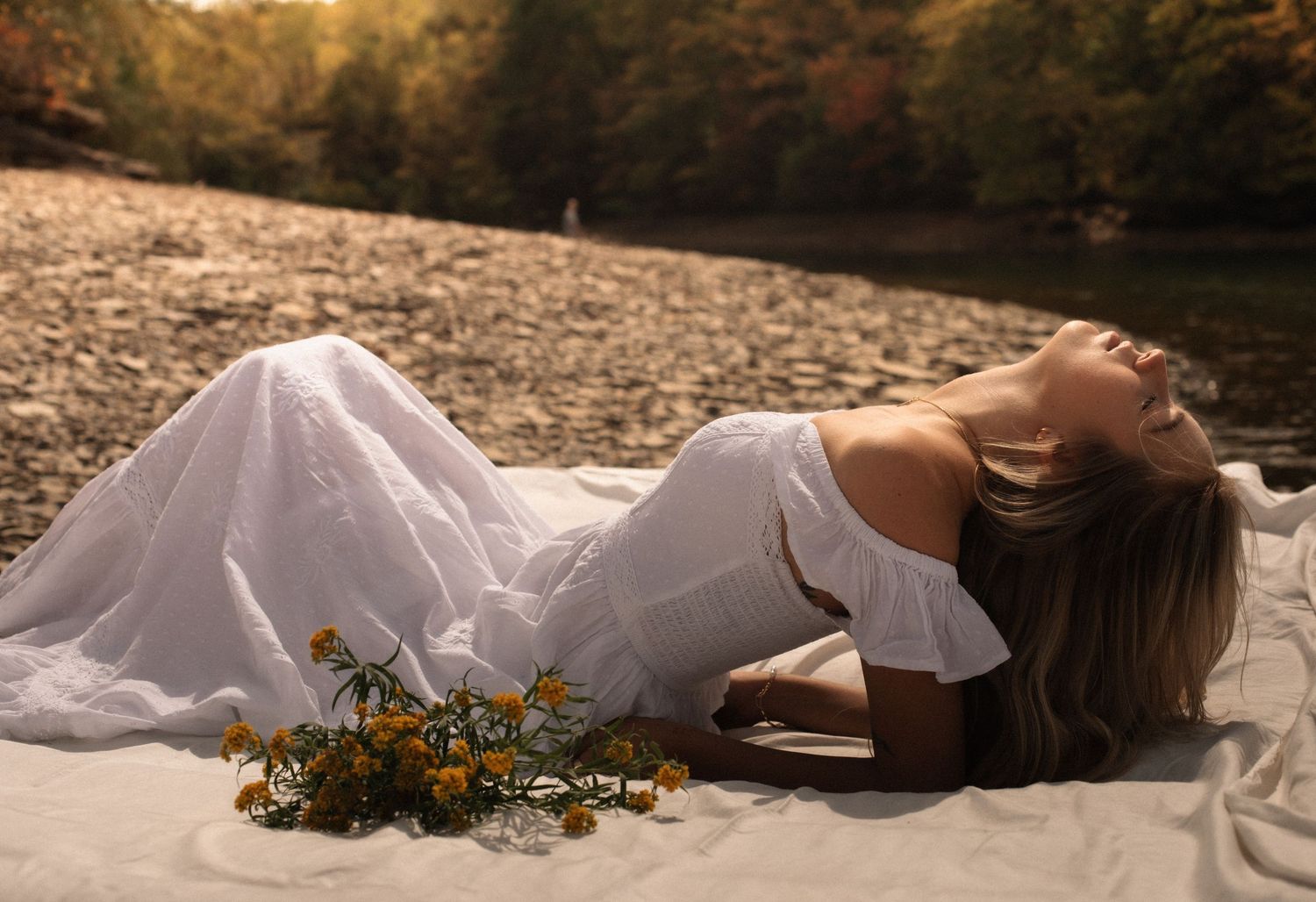 A woman in a white off-shoulder dress lies on a blanket by a rocky riverbank with yellow wildflowers nearby.