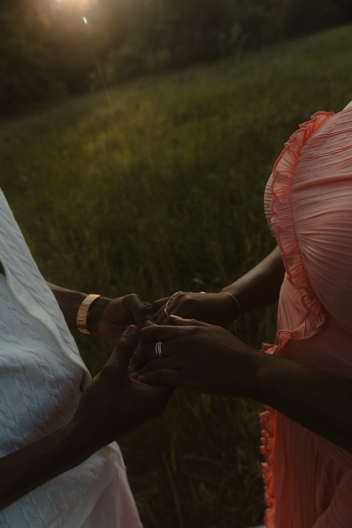 Couple holding hands at sunset in a green field, wearing white and coral outfits with gold jewelry.