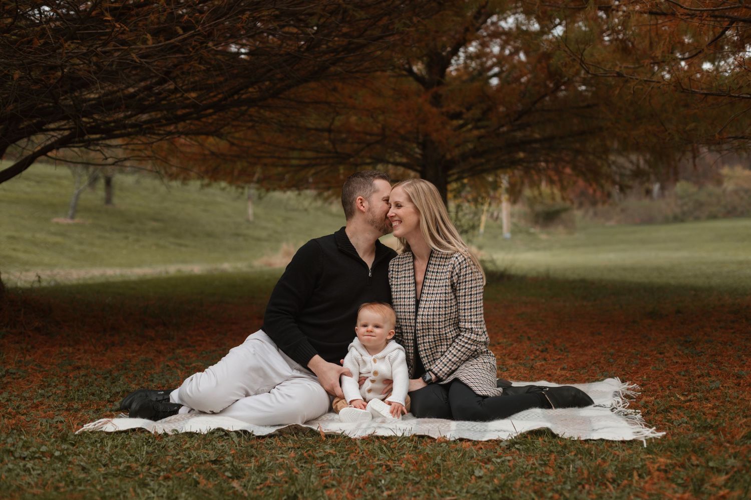 Couple sitting together on white blanket surrounded by autumn trees in a park setting.
