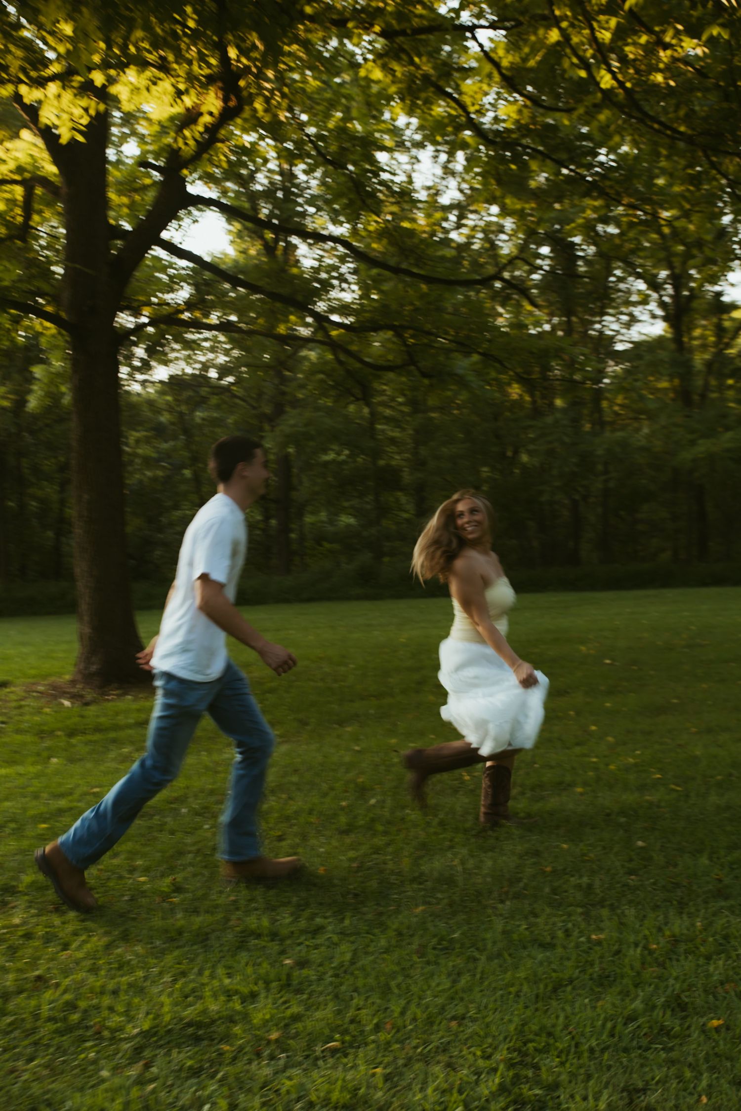 A joyful couple runs through a lush green park, woman in white dress laughing as man chases her at golden hour.