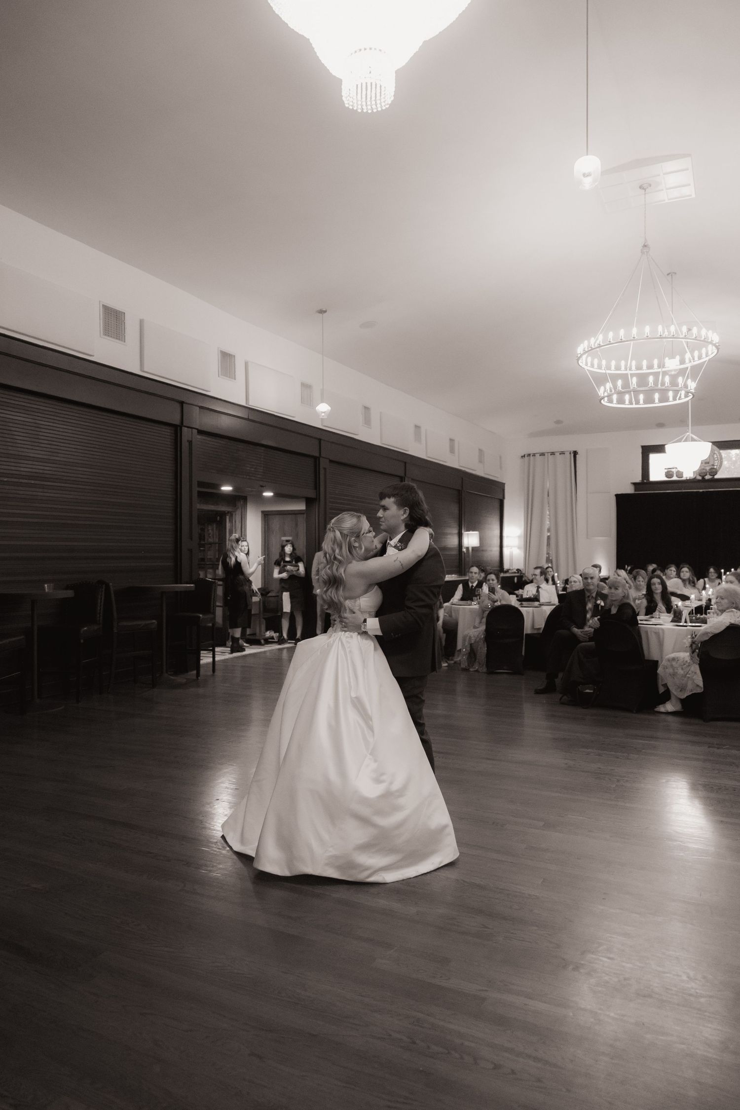 Black and white photo of a bride and groom sharing their first dance in an elegant ballroom with chandelier lighting.