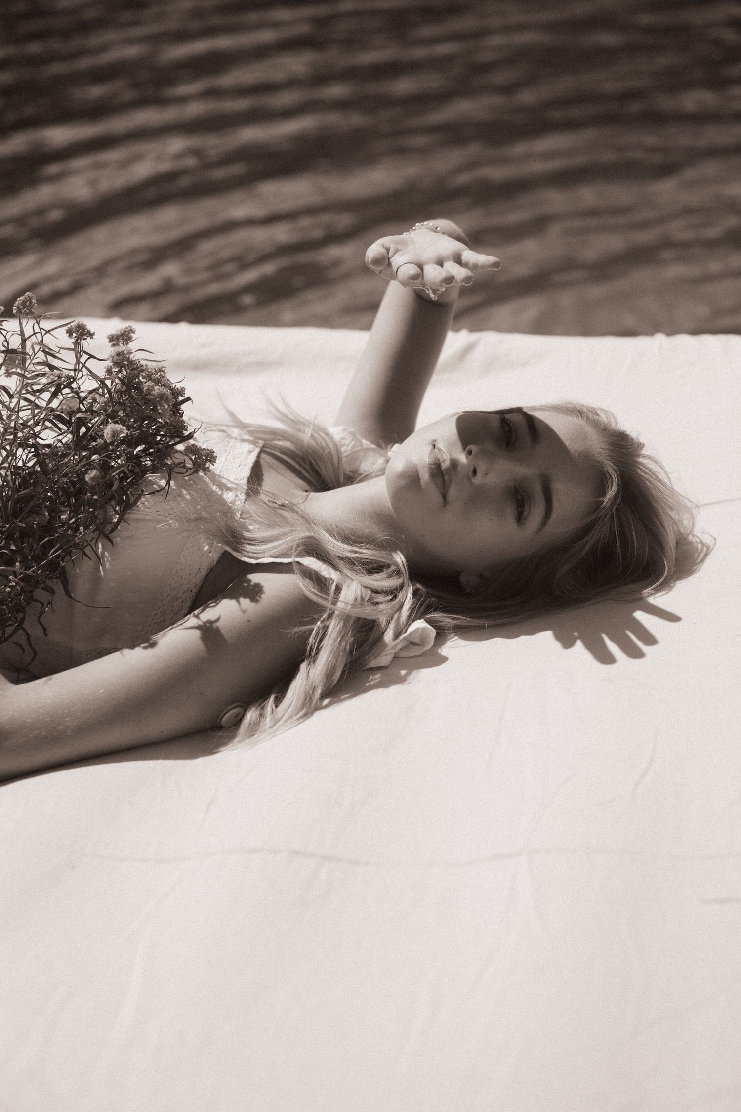 Black and white photo of a young woman lying on white fabric holding wildflowers, with sunlight casting shadows.