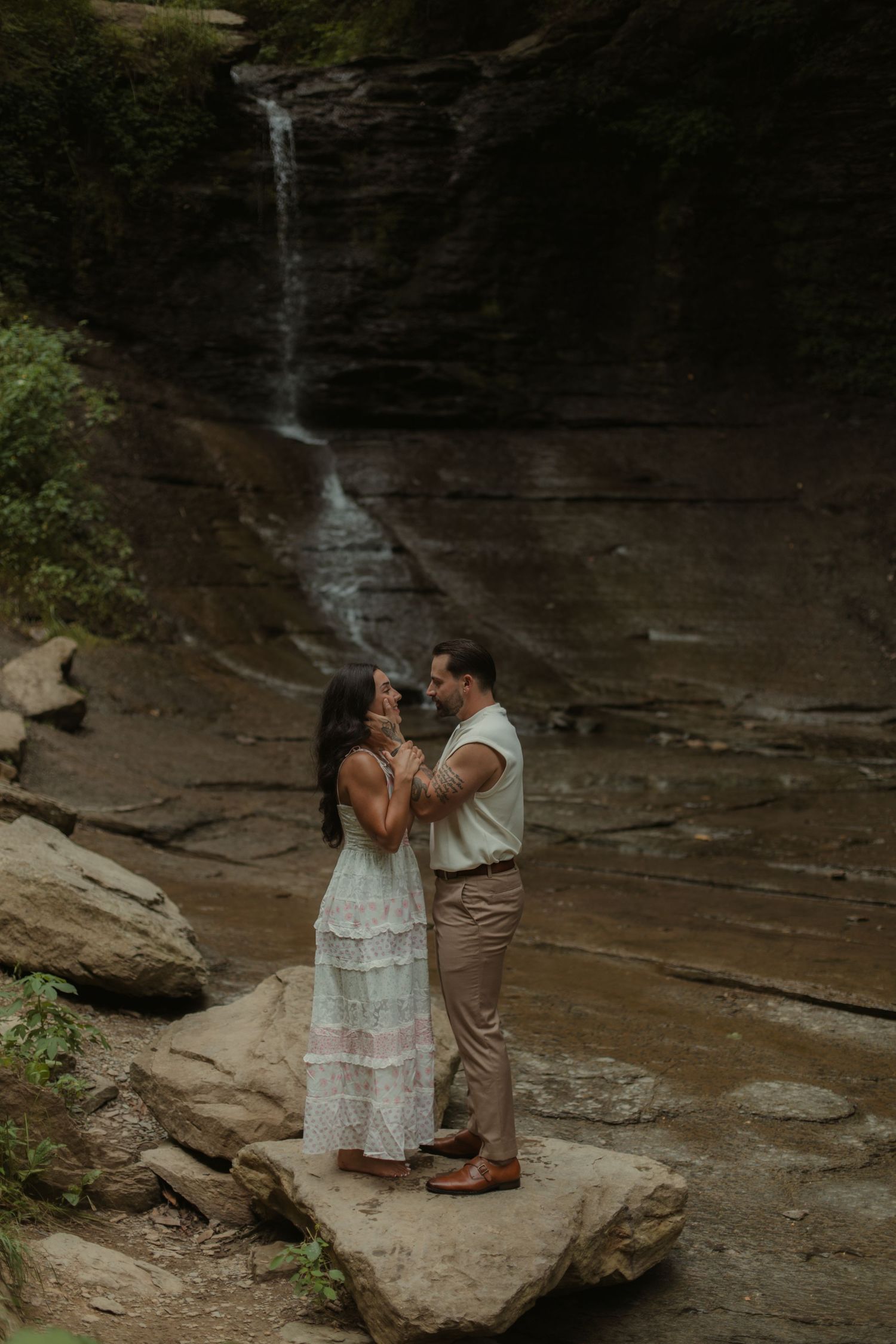 Couple standing on rocks in front of a waterfall in a lush gorge, woman in white dress, man in tan pants.