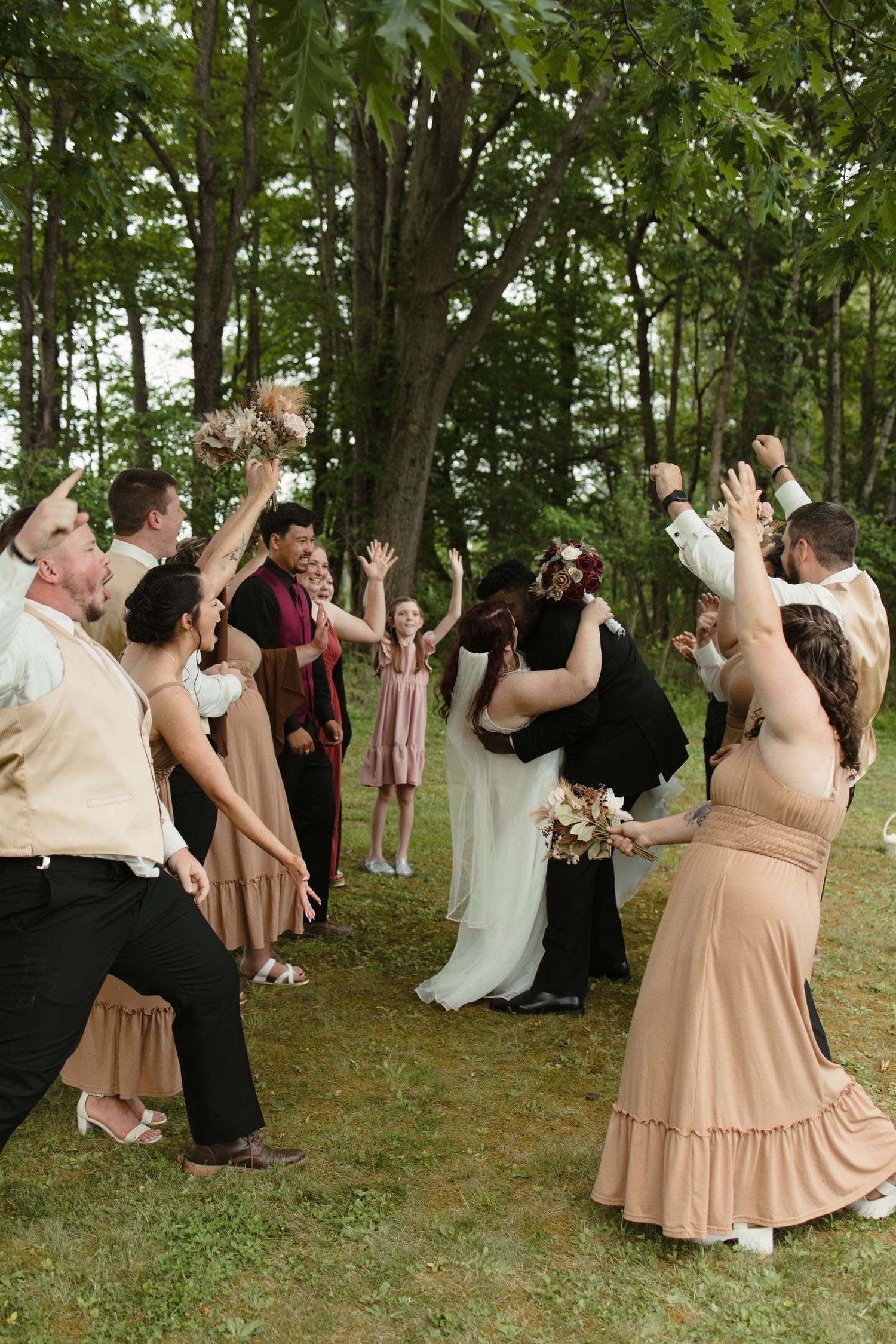 Wedding party celebrates outdoors among trees, with bride and groom kissing while guests cheer with raised arms.
