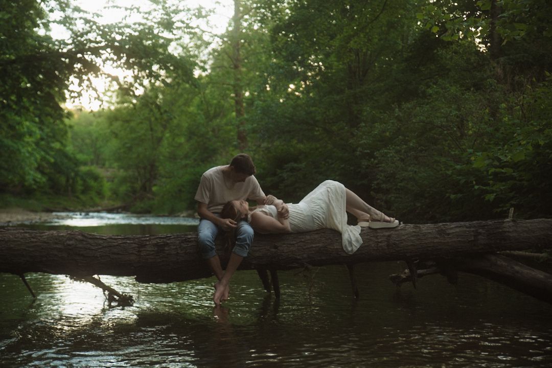 Couple sitting on a fallen log over a peaceful river surrounded by lush green forest trees.
