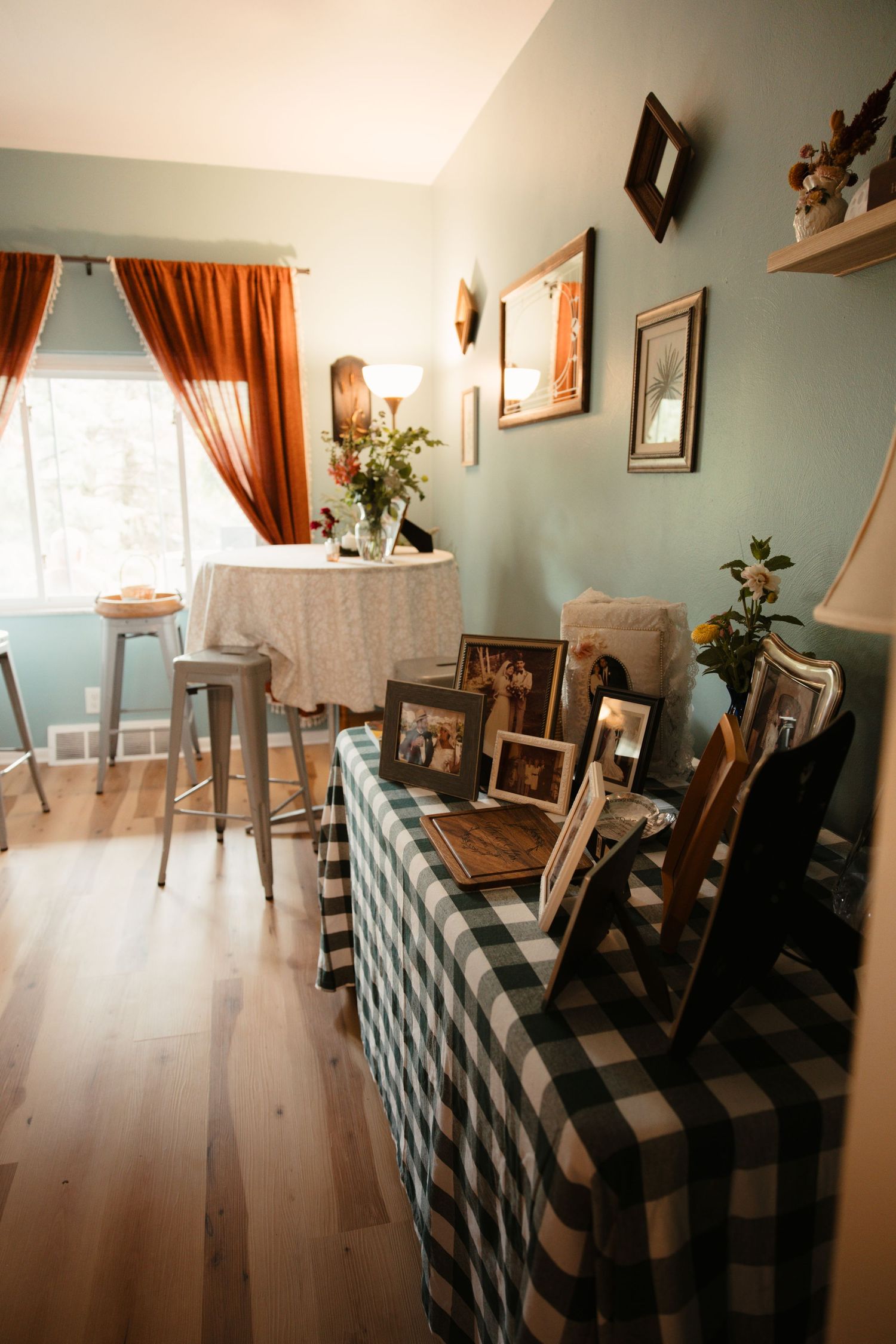 Cozy dining room with plaid tablecloth, framed photos, rust curtains, and sage green walls with hardwood floors.