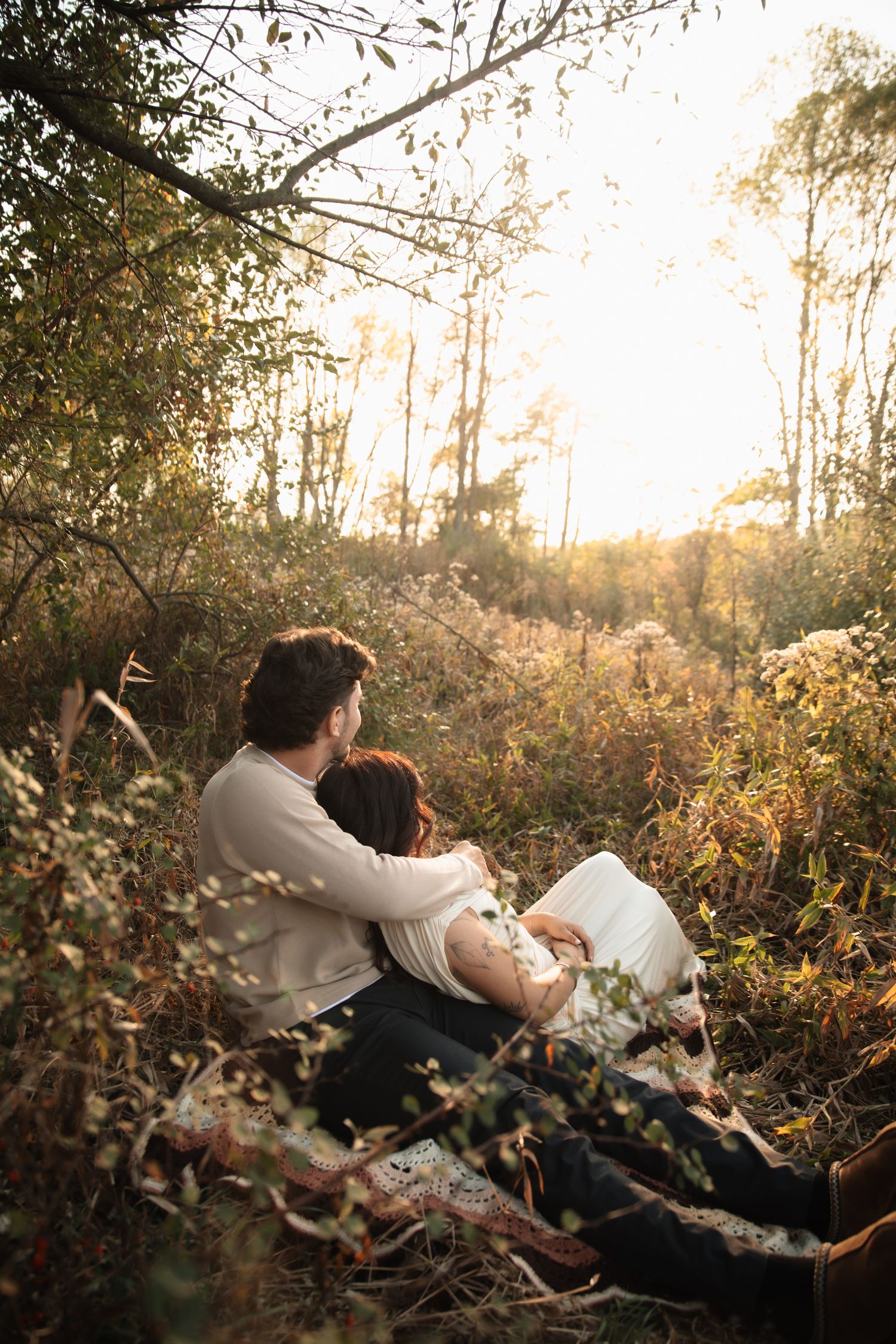 Couple sitting together in golden hour sunlight surrounded by wild grass and autumn trees in a romantic outdoor setting.