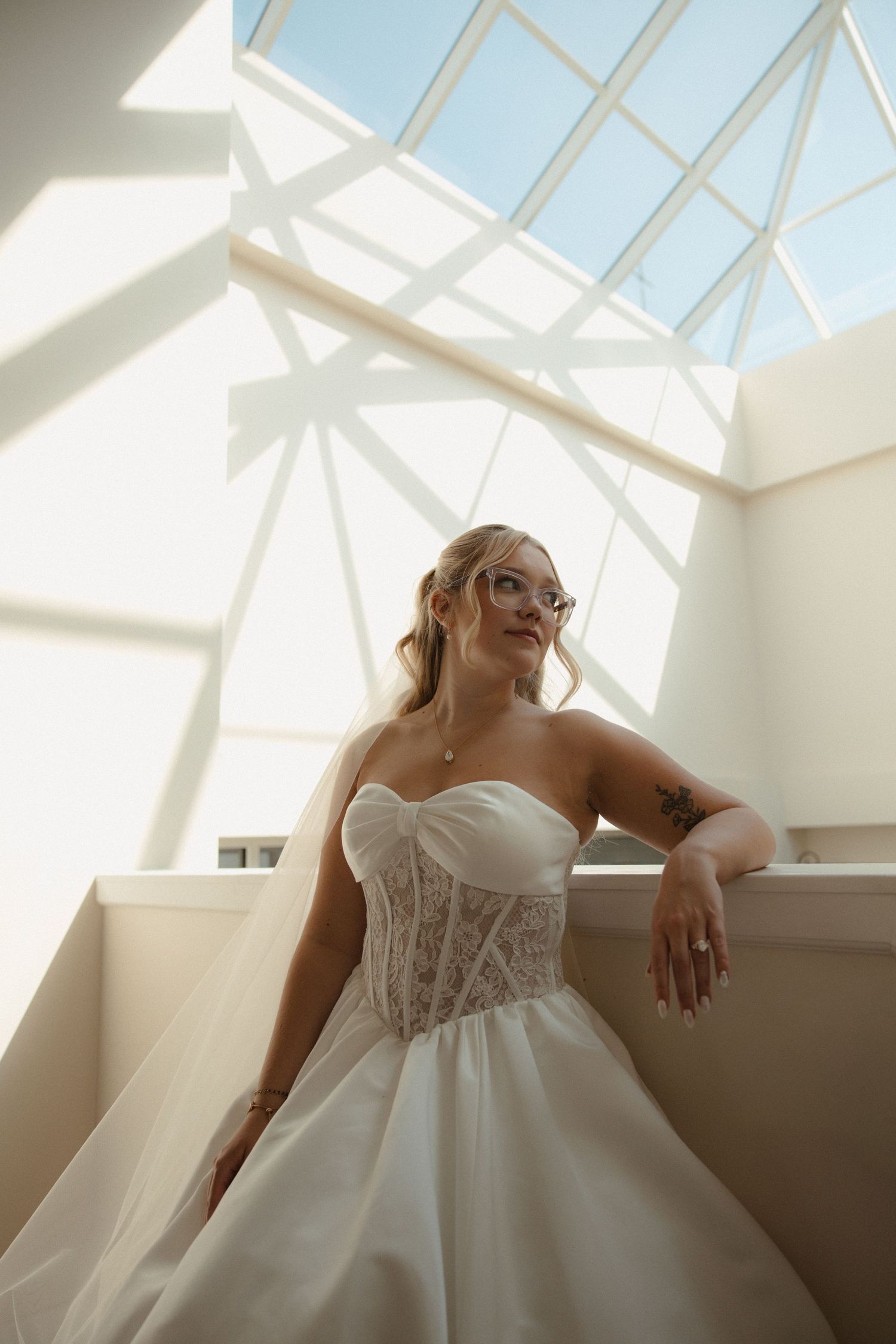 Bride in strapless lace corset ball gown poses under geometric skylight in bright airy venue.