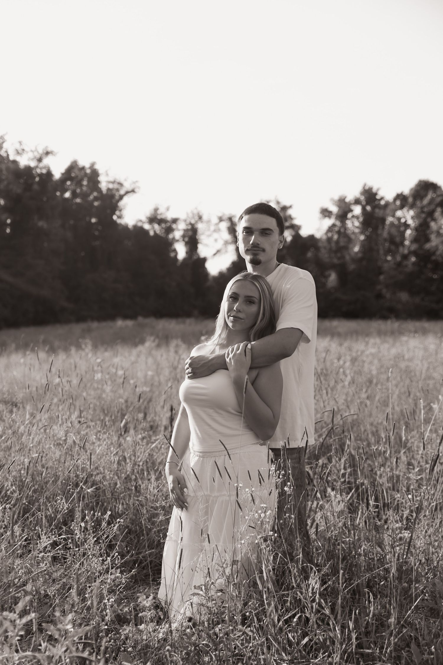 Black and white photo of a couple embracing in a field, man hugging woman from behind among tall grass with trees behind.