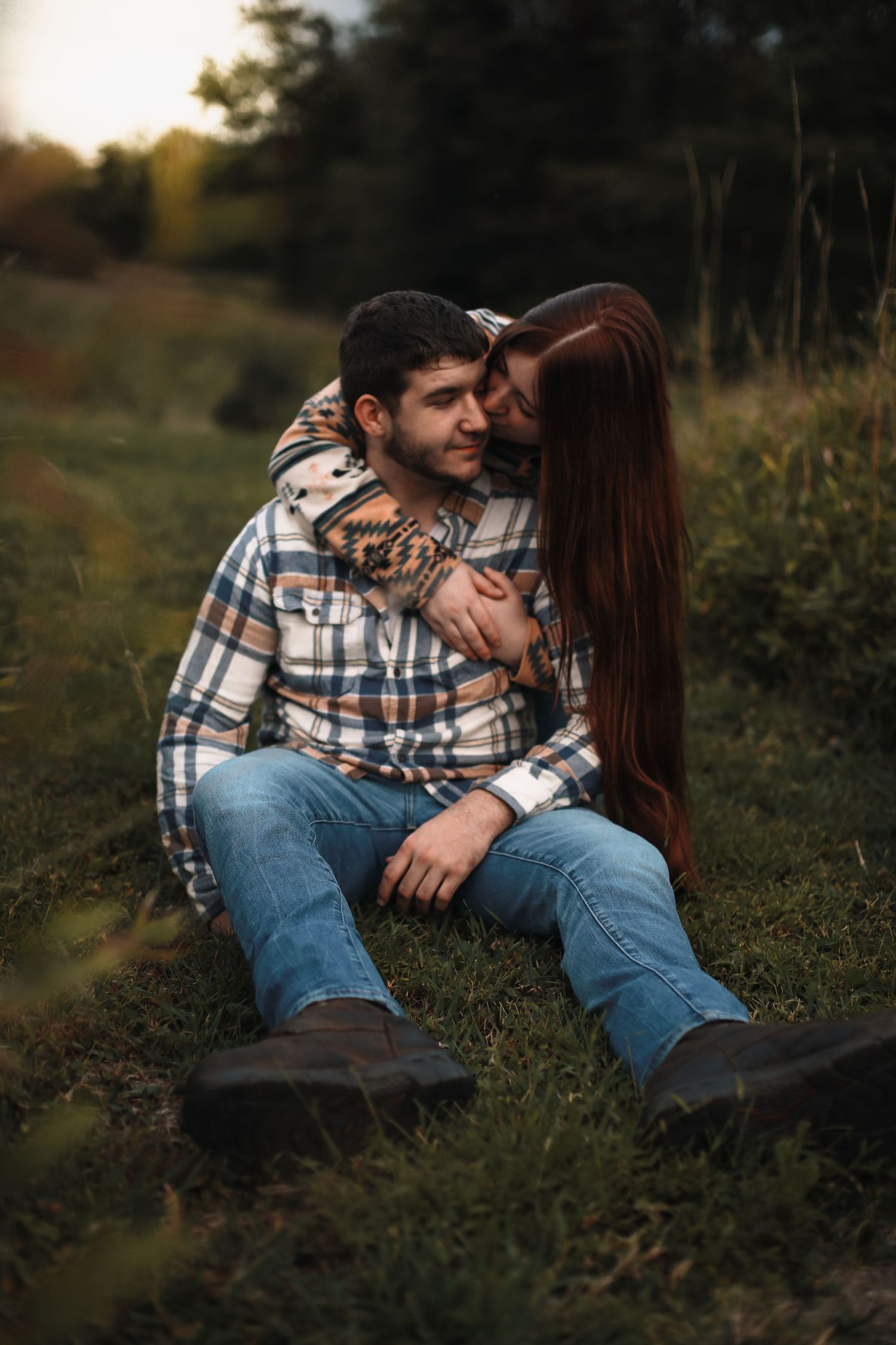 A romantic couple sits together on grassy ground at sunset wearing casual attire in a natural outdoor setting.