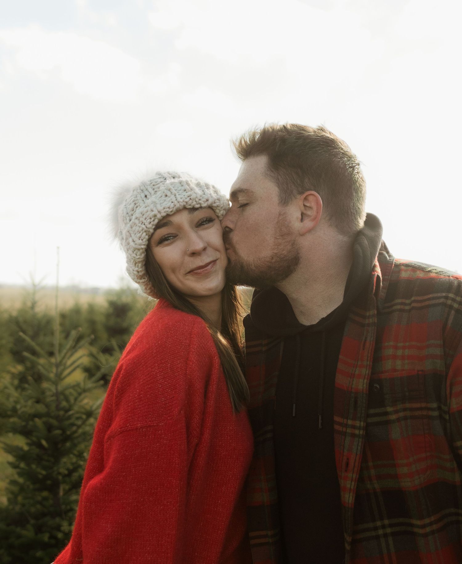 A couple shares a tender moment among evergreen trees on a cloudy day, wearing winter attire including a knit hat and plaid jacket.