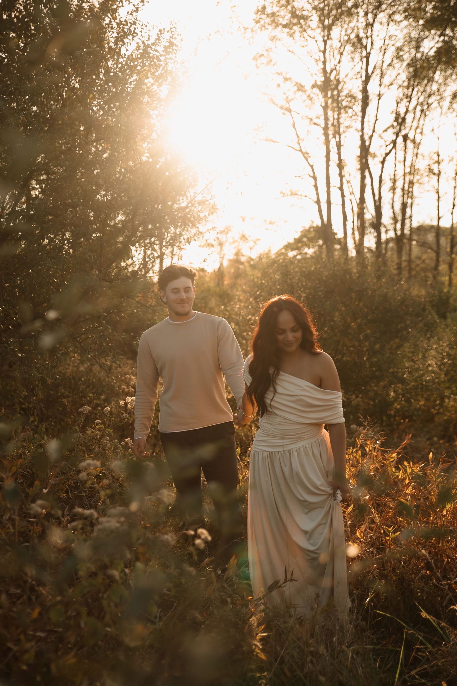 A couple walks through a sunlit meadow at golden hour with tall trees and wildflowers in the background.