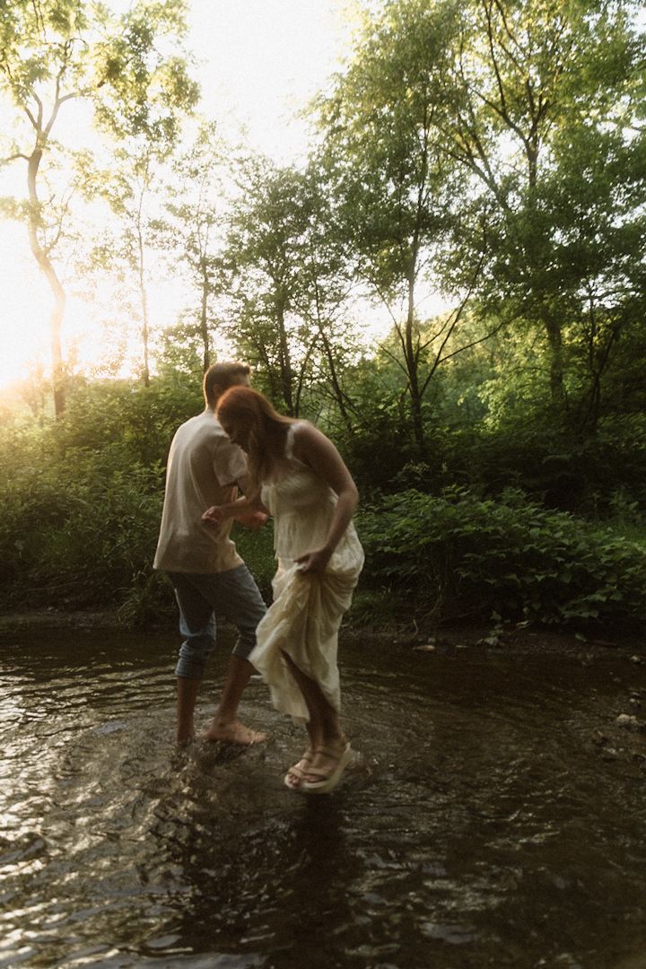 Couple wading barefoot in a sunlit forest stream, woman in white dress, golden hour light filtering through green trees.