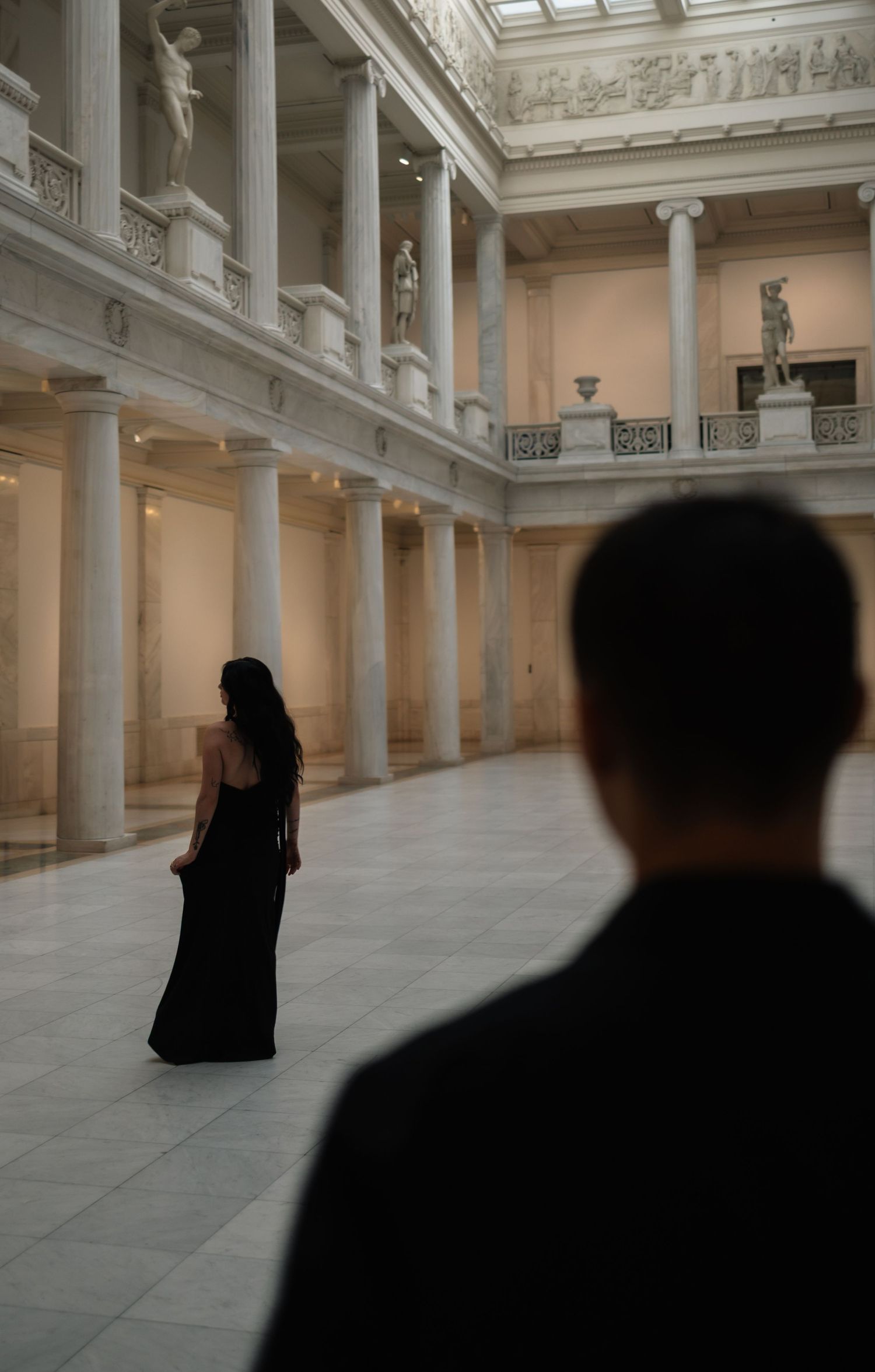 Woman in black gown stands in neoclassical museum hall with columns and sculptures, man in foreground.
