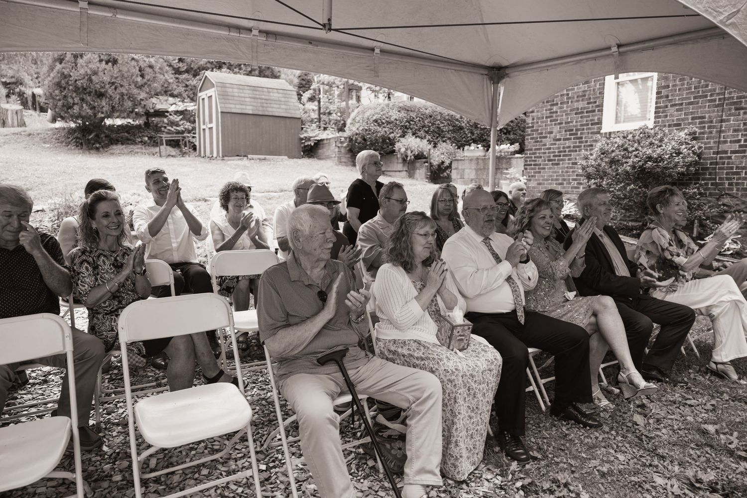 Black and white photo of a crowd of people clapping and seated outdoors under a tent at a gathering.