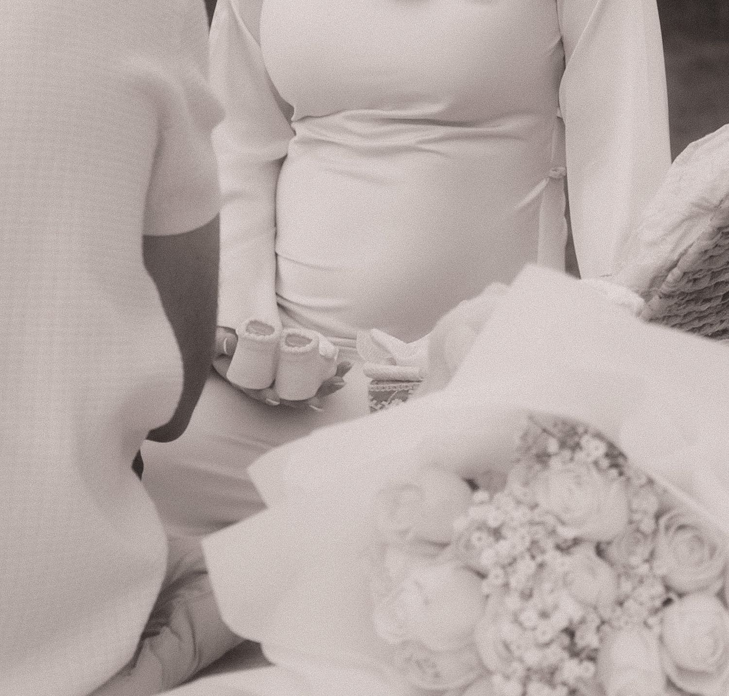 Black and white photo of a pregnant bride in white dress holding a bouquet of roses at a wedding ceremony.