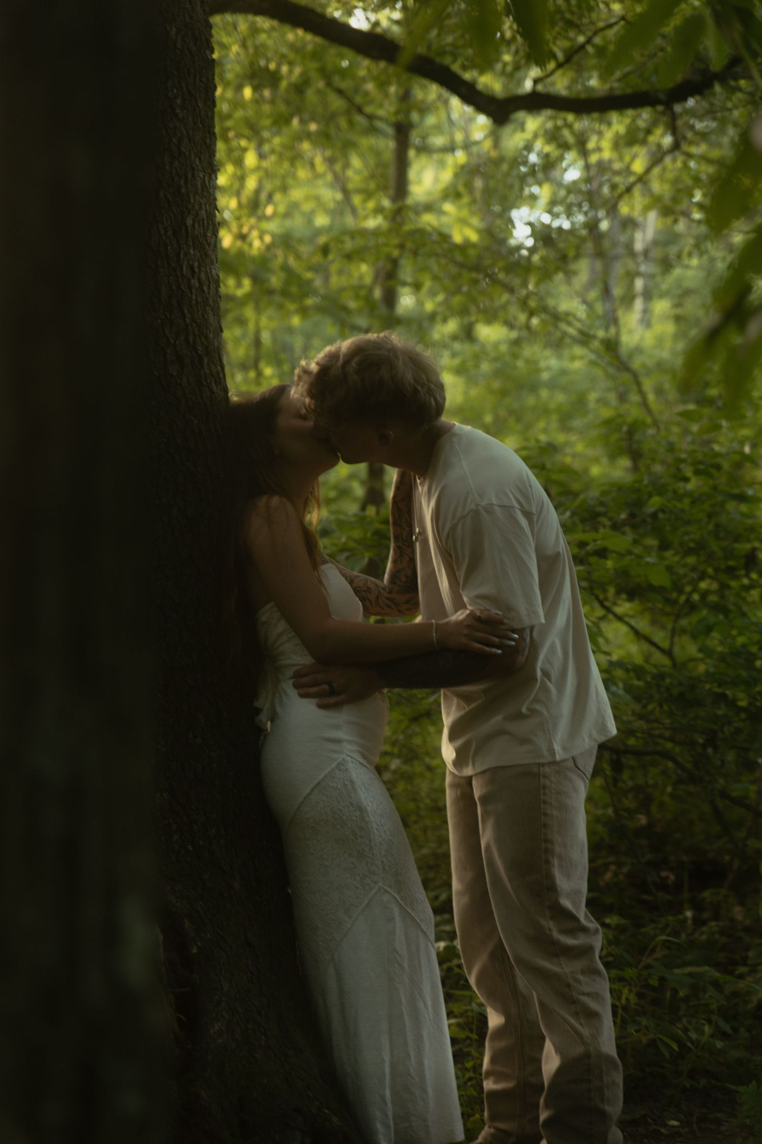 Couple sharing tender kiss leaning against tree in lush green forest setting.