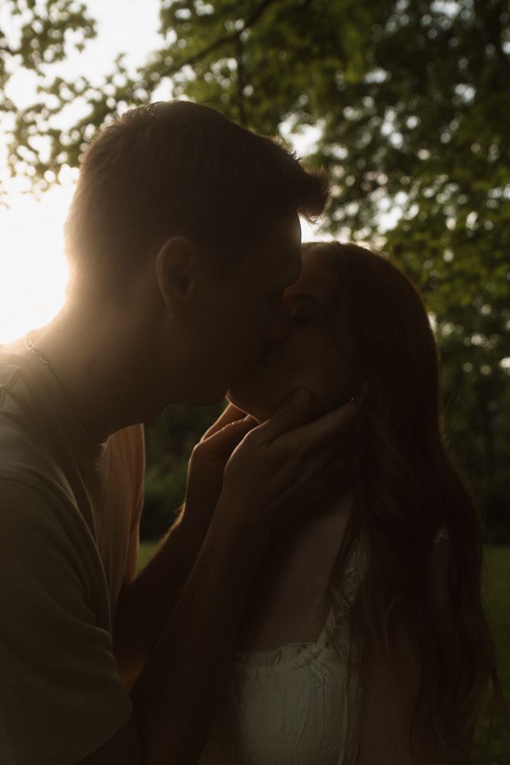 Romantic couple sharing a tender kiss at golden hour, backlit by warm sunset light in a lush green garden.