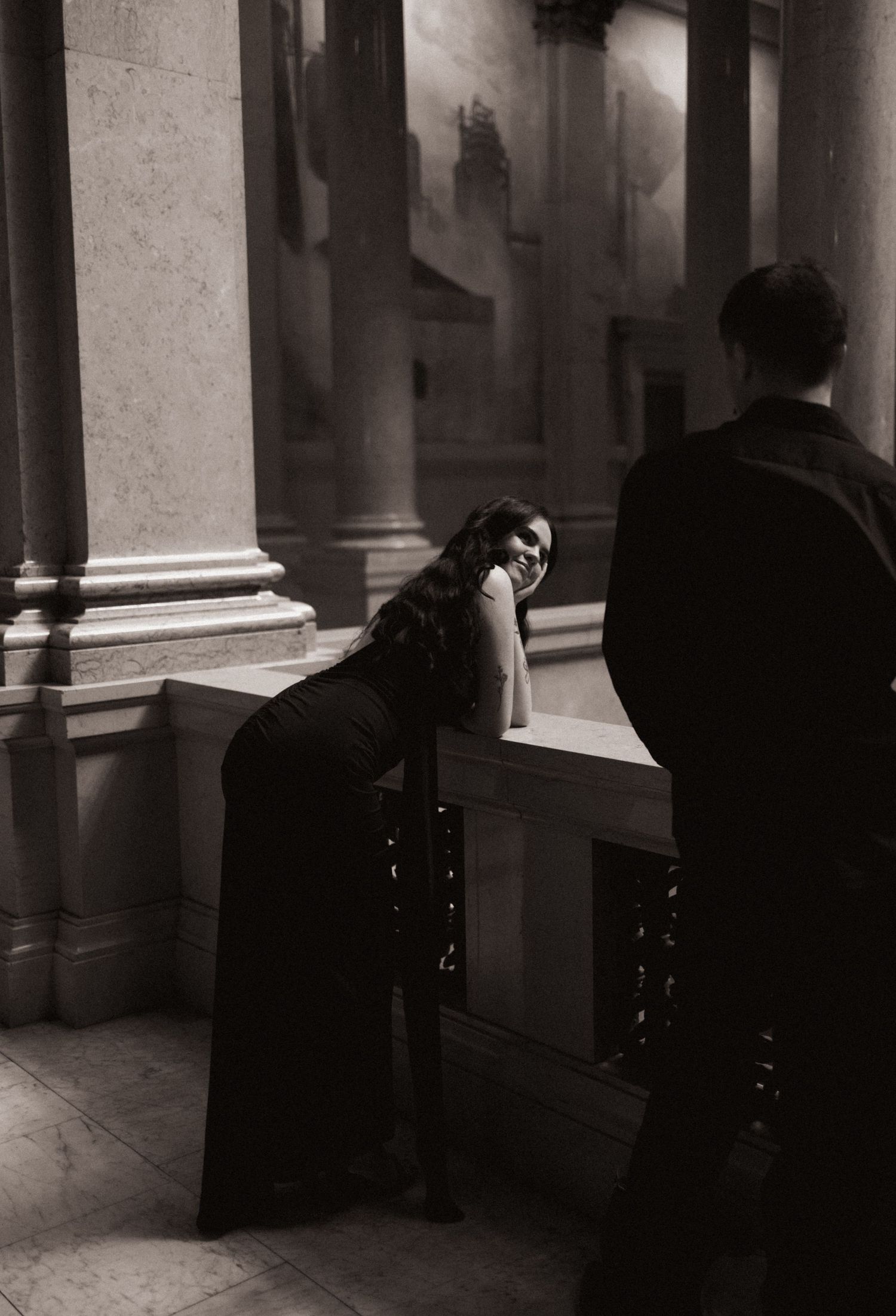 Woman in black dress leaning on railing smiling at man in moody architectural setting.