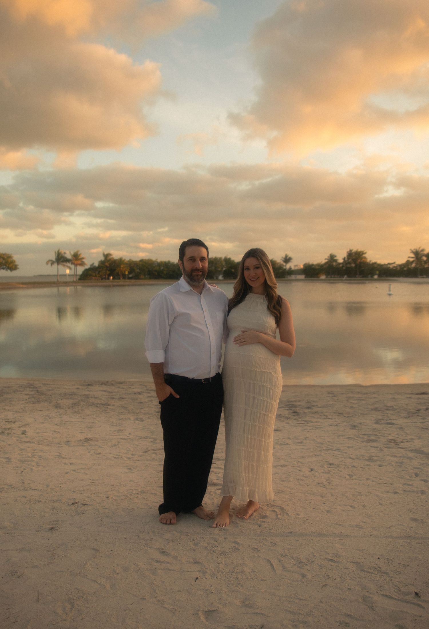 Couple poses together on a sandy beach at sunset with palm trees and calm waters in the background.