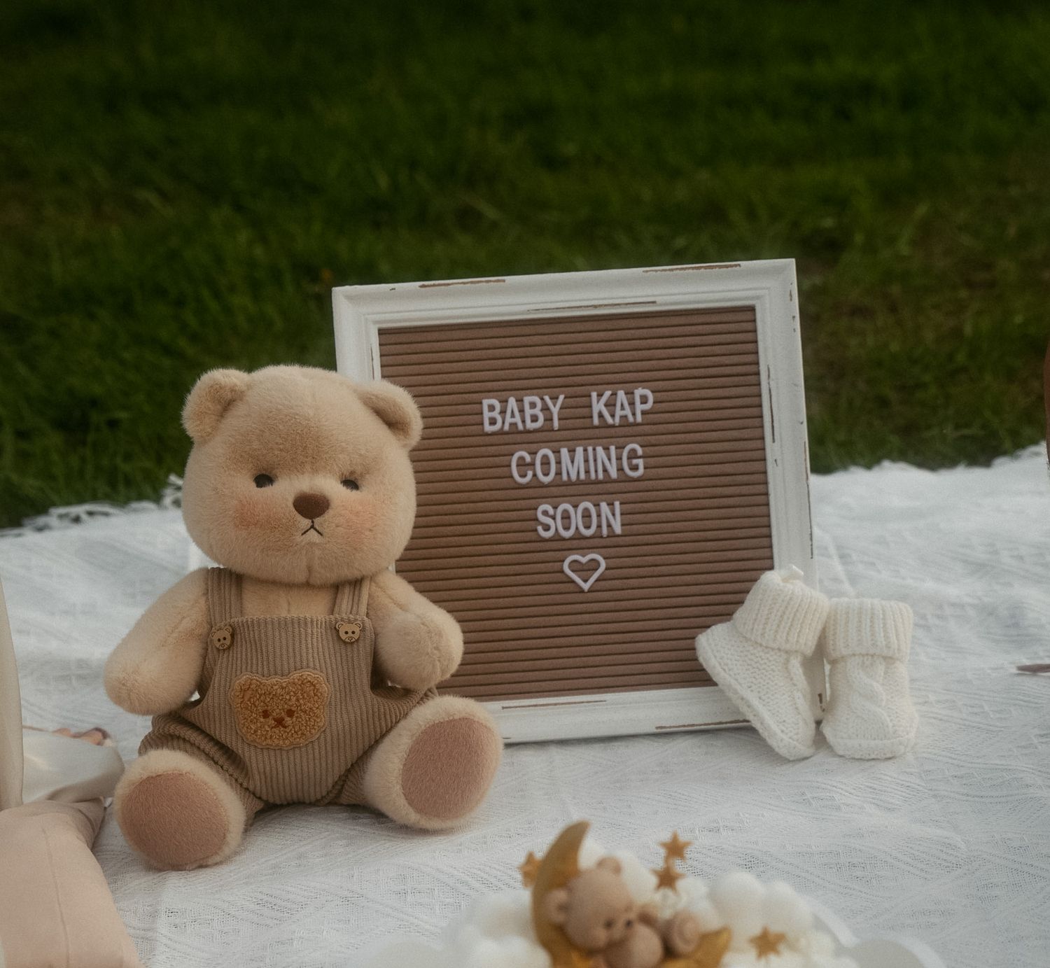 Teddy bear beside a letter board reading Baby KAP Coming Soon on a white blanket outdoors.