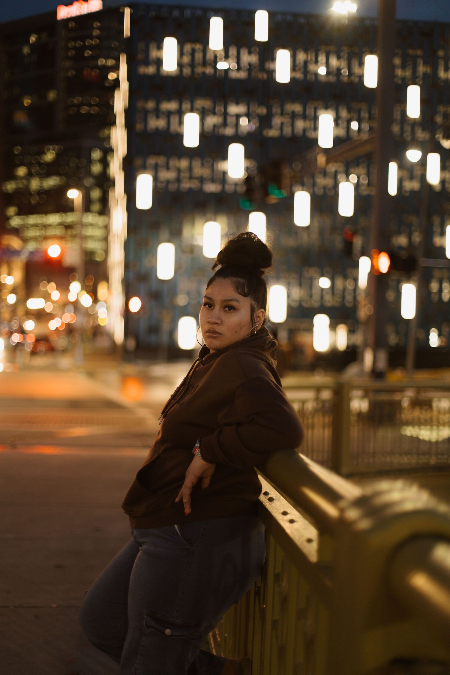 Young woman in brown hoodie and jeans leans on railing at night with city lights bokeh background.