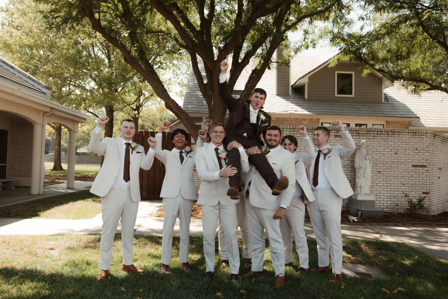 Groomsmen in white suits celebrate outdoors, lifting the groom in a joyful wedding party photo near a tree.