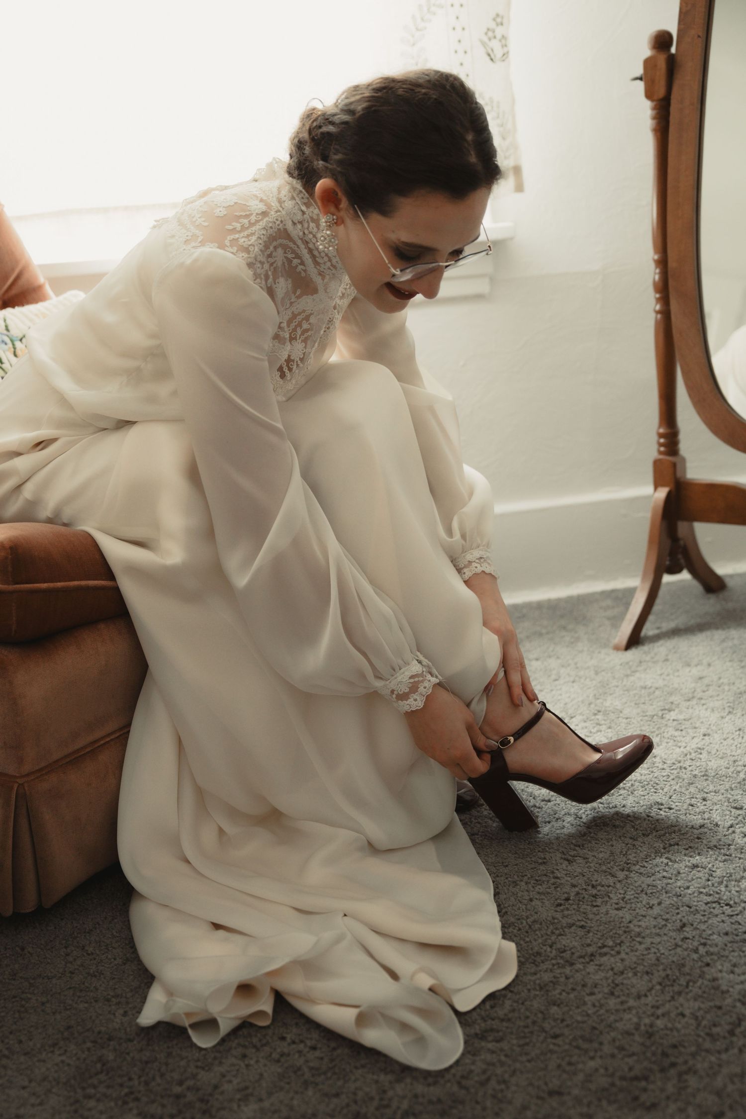 Bride in white lace gown sitting on chair fastening brown Mary Jane heels, preparing for wedding day.