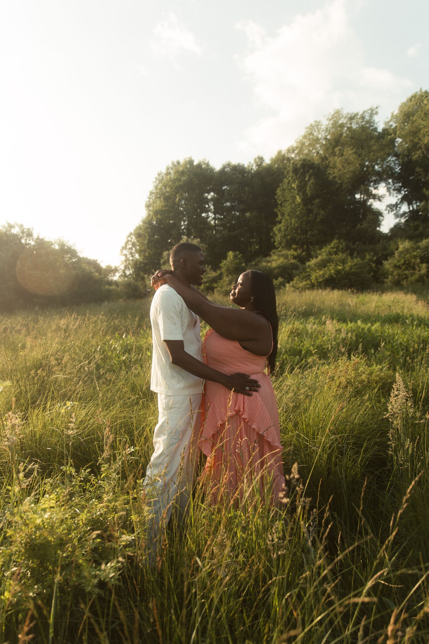 A couple embraces in a sunlit meadow, the woman in a pink dress and man in white, surrounded by tall grass and trees.