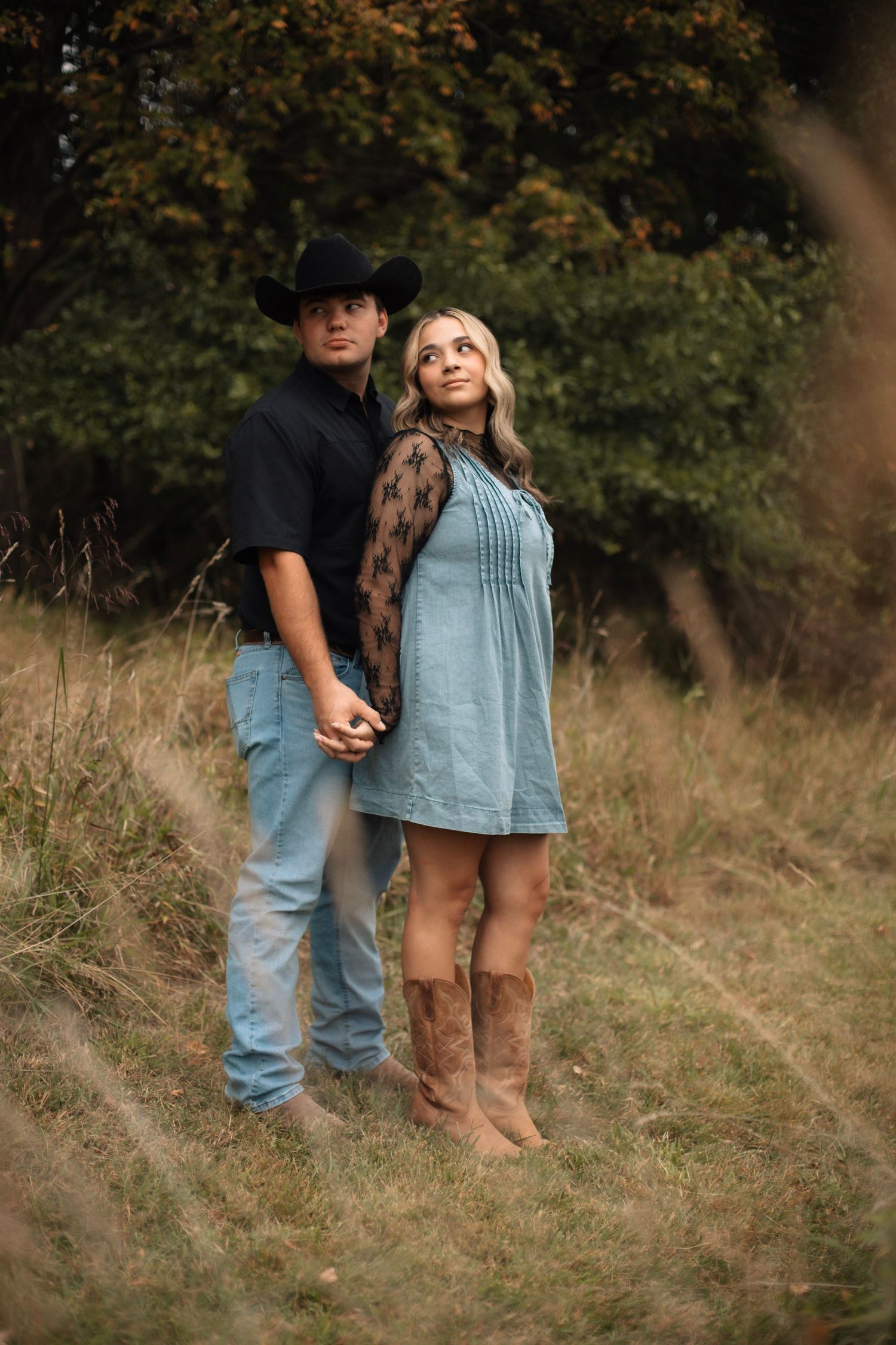 A couple in country western attire holds hands in a field, man in black cowboy hat, woman in blue dress and cowboy boots.