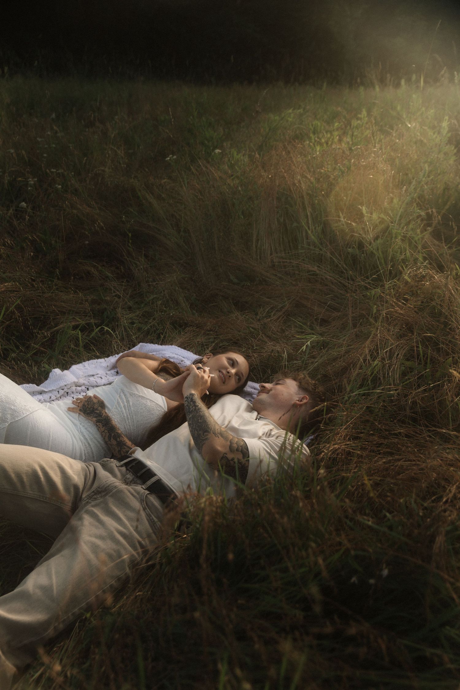 Romantic couple lying in tall grass field, holding hands in warm golden sunlight.
