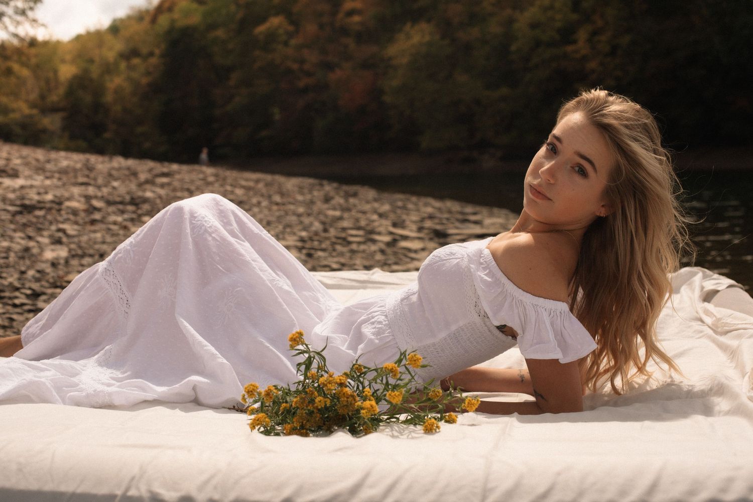 Romantic outdoor portrait on white blanket with wildflowers at sunset by rocky riverbank in autumn.