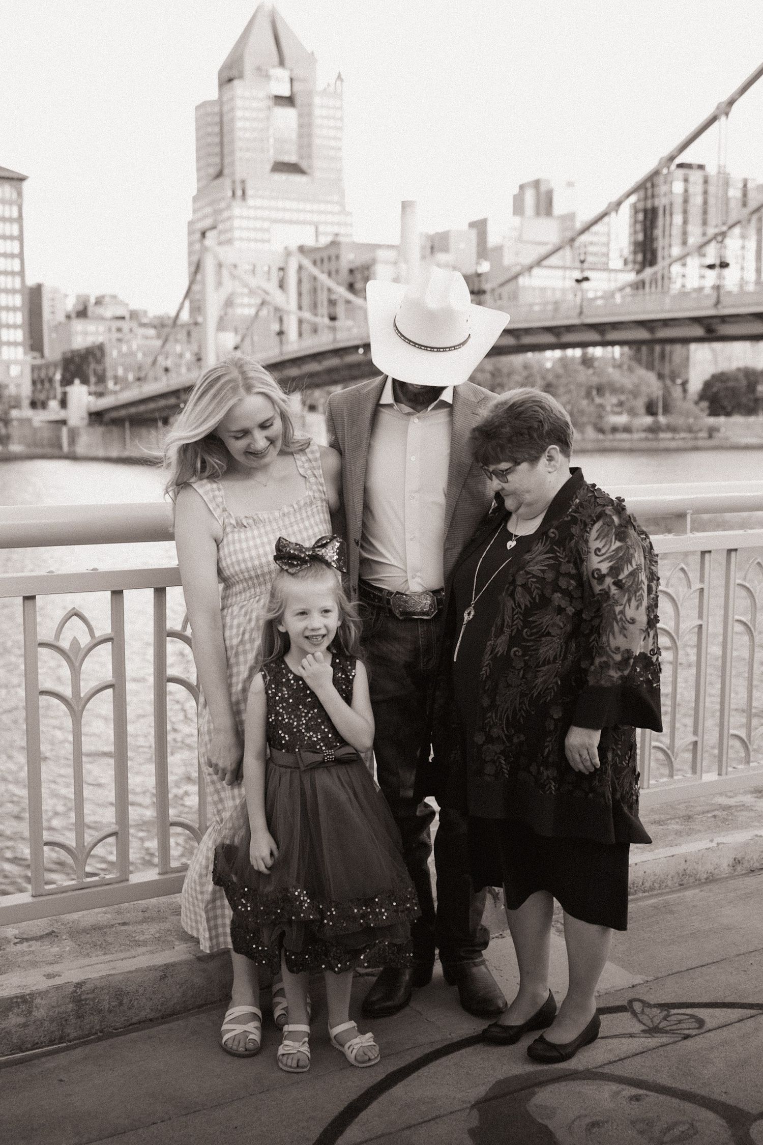 Black and white family photo of four people posing on a bridge in Pittsburgh, PA with city skyline in background.