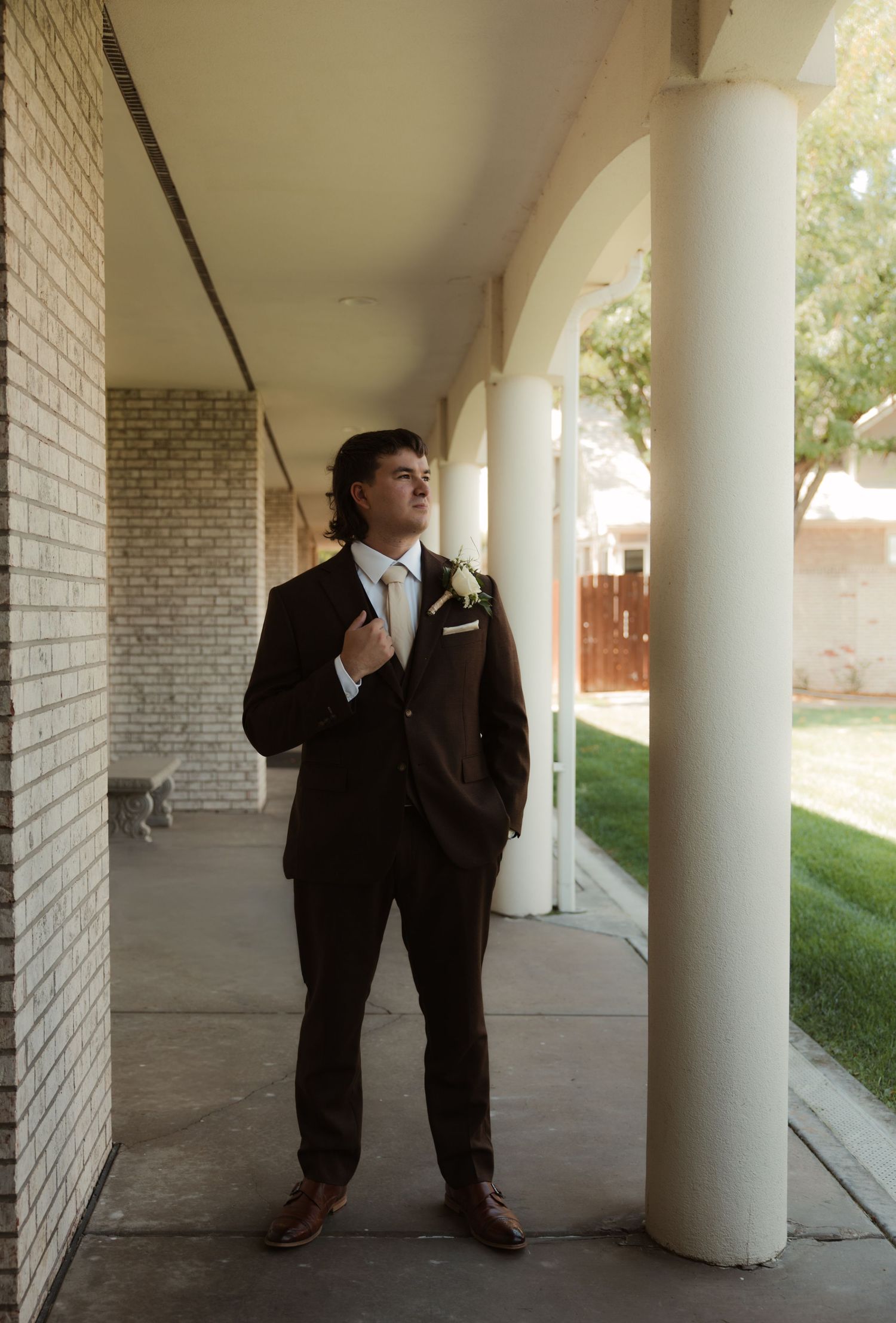 A groom in a brown suit with boutonniere adjusts his tie while standing under arched columns at a wedding venue.