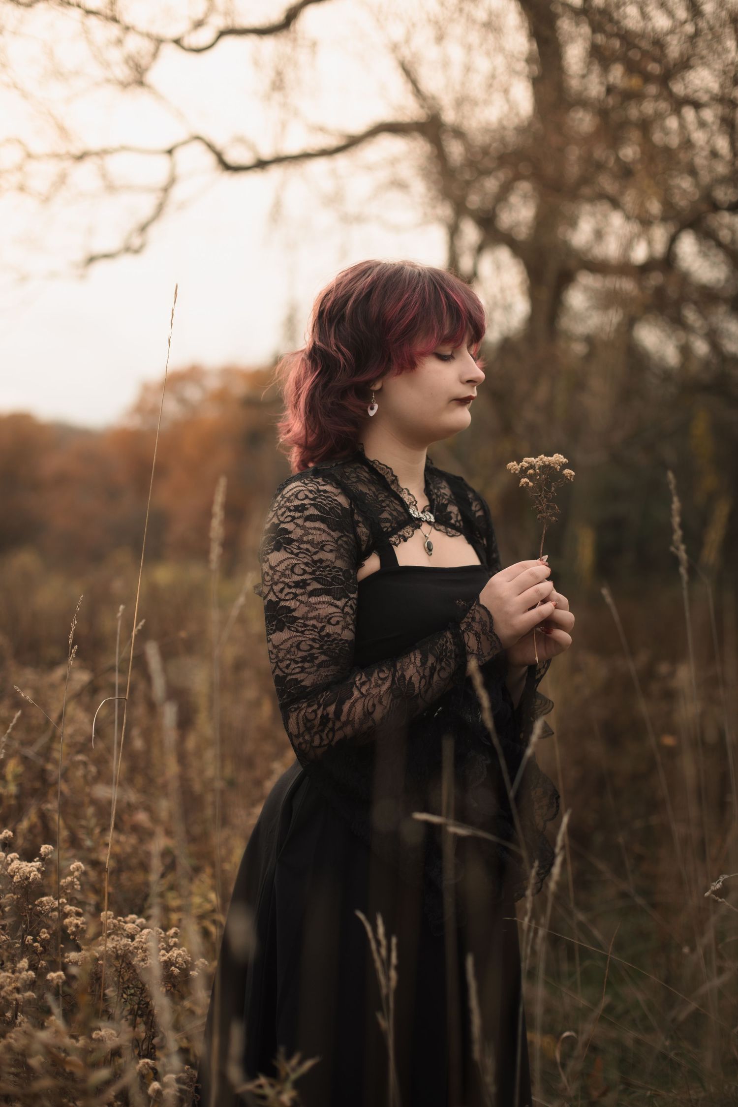 A person with burgundy hair wearing a dark coat holds a dried plant in an autumn field at dusk.