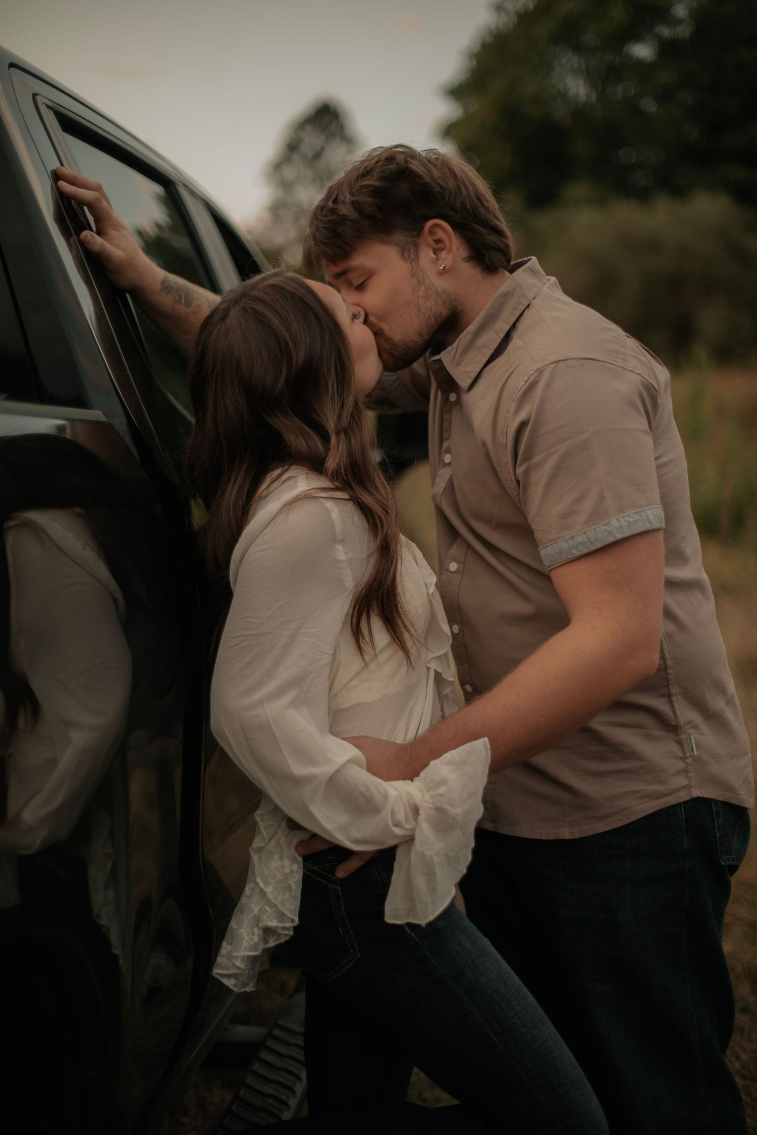 A romantic couple embraces against a car in warm evening light wearing casual attire.