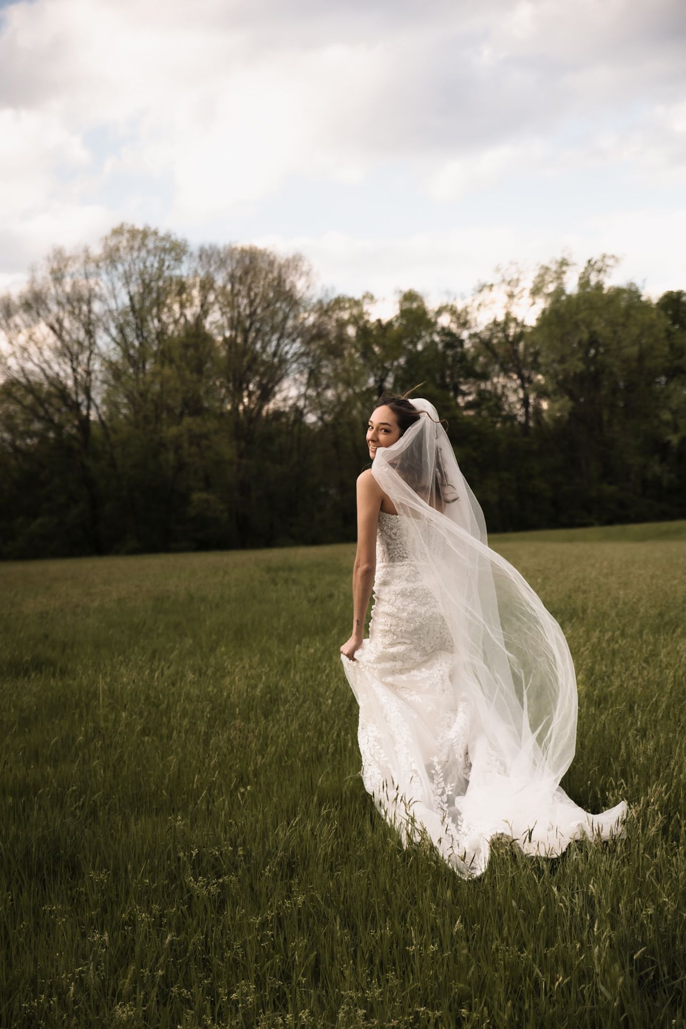 Bride in lace wedding dress with long flowing veil blowing in wind stands in green field near trees.
