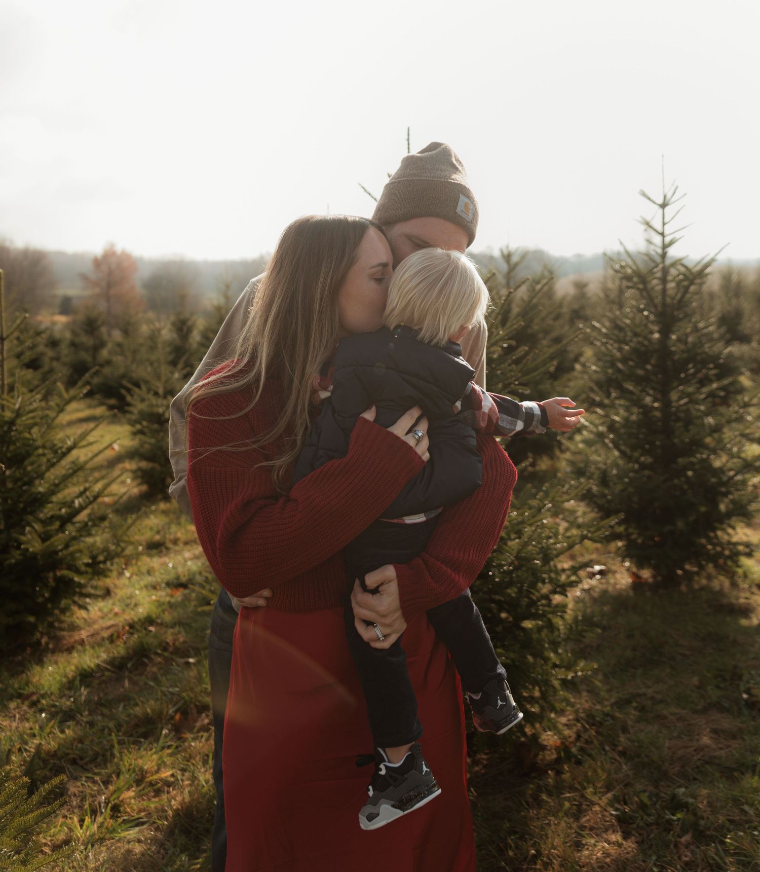 A couple embraces while standing in a Christmas tree farm during golden hour, creating a warm and intimate moment.
