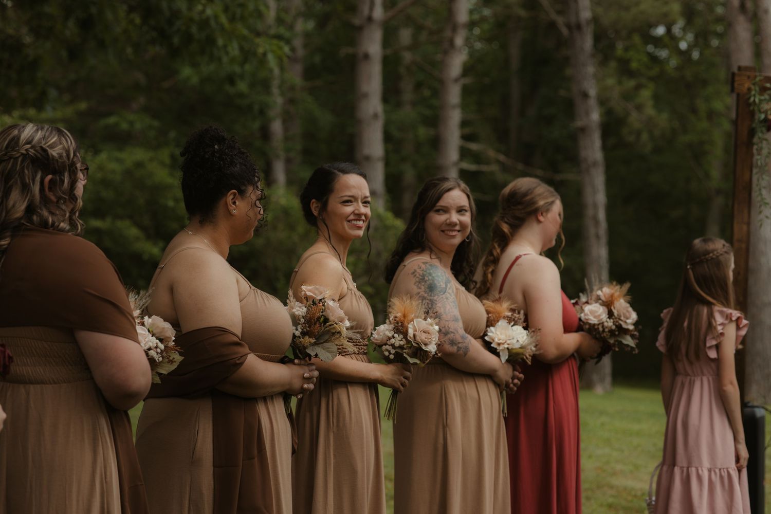 Bridesmaids in tan and red dresses holding floral bouquets stand outdoors at a woodland wedding ceremony.
