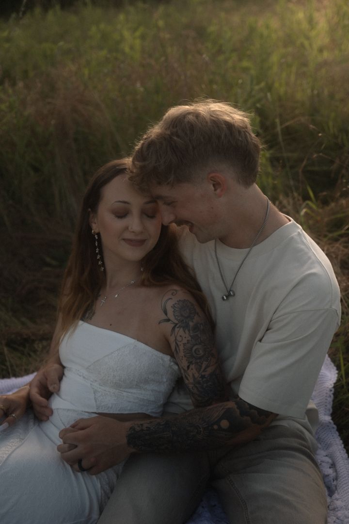 A couple sits in a sunlit field, the tattooed man nuzzling the smiling woman in a white dress.