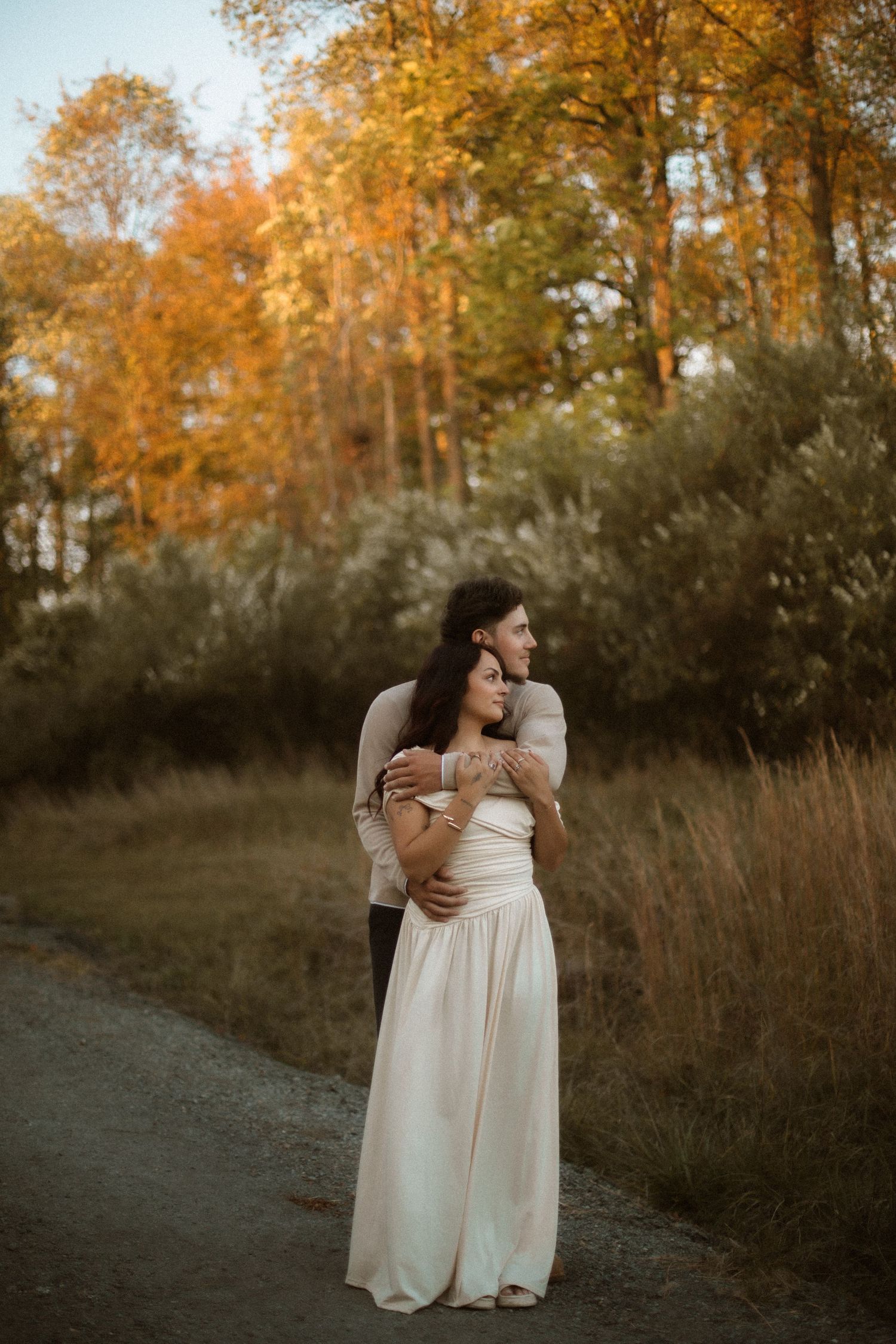 Couple embracing on a path surrounded by autumn foliage, woman in white gown, romantic fall engagement photo.