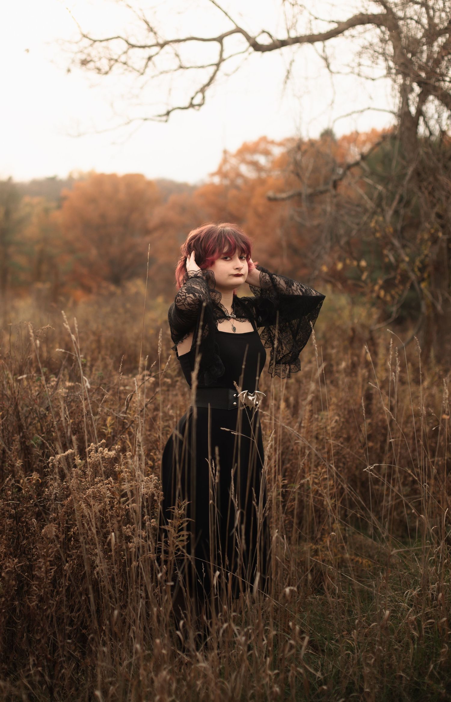 Woman in black gothic dress stands in autumn field with red hair, surrounded by dried grasses and colorful fall foliage.
