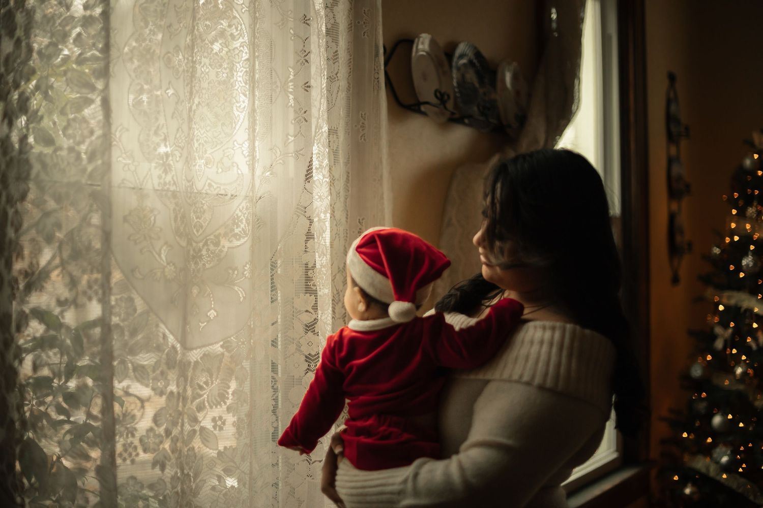 Mother holding baby in Santa hat looking out window with Christmas tree in background.