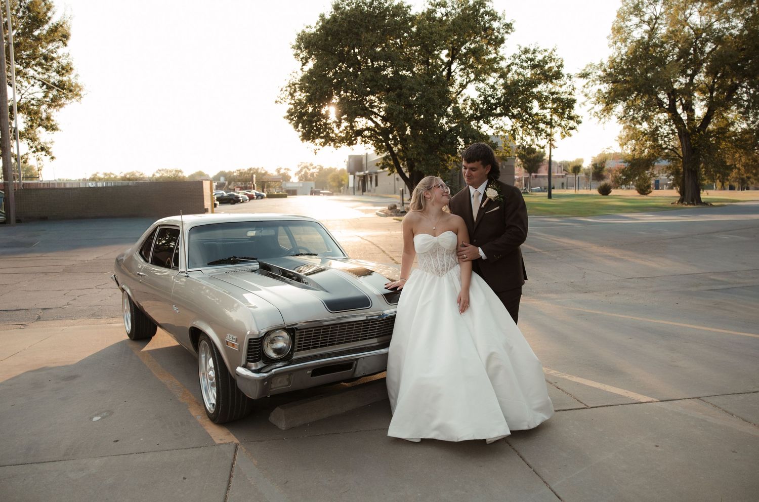 Bride and groom pose beside a classic silver muscle car at sunset on their wedding day.